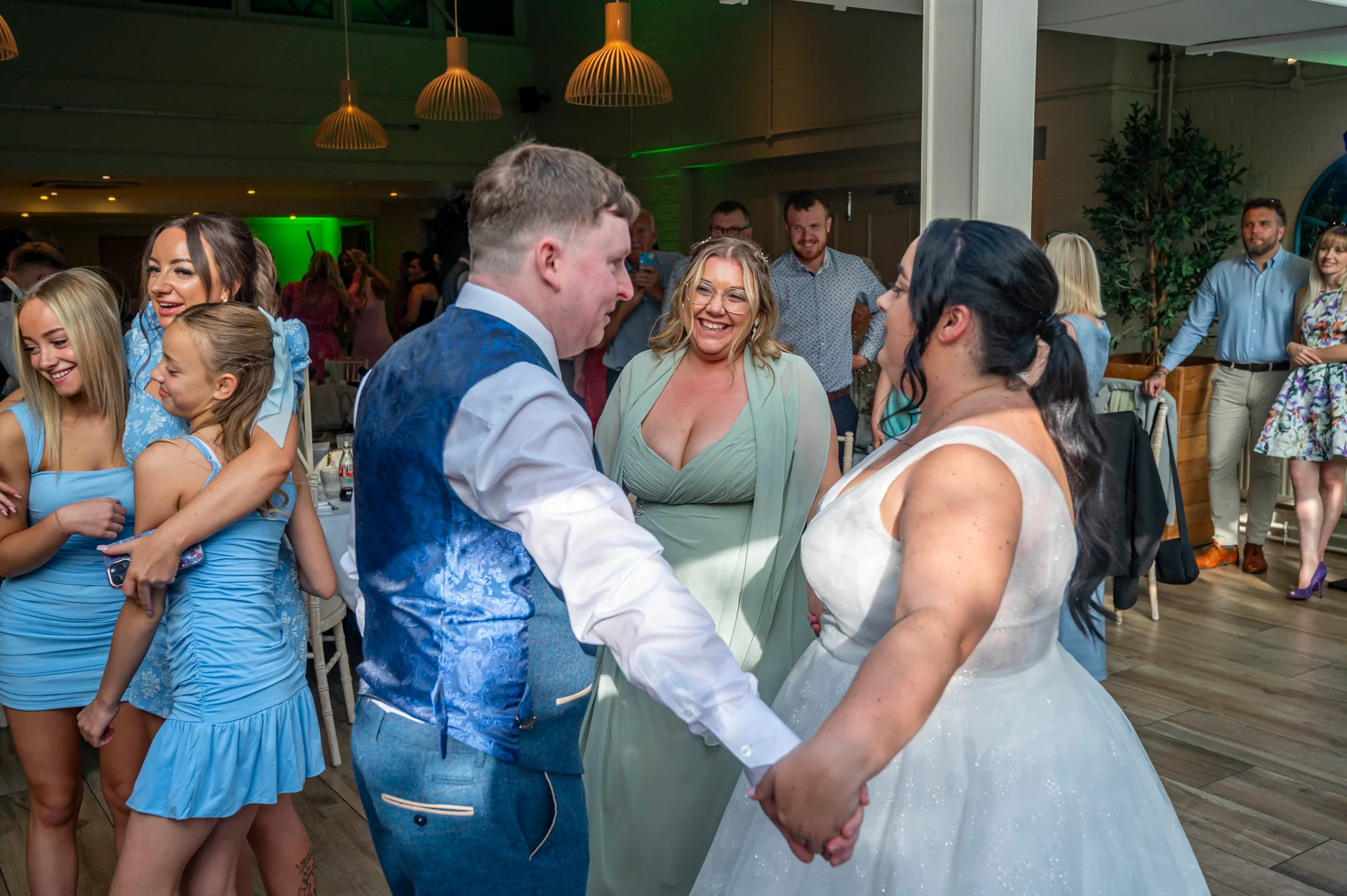 The bride and groom share a joyful first dance while surrounded by smiling family members and guests inside a reception venue.