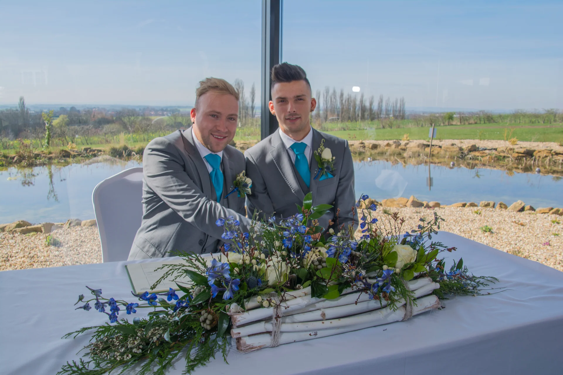 Two grooms in matching grey suits and bright blue ties sit together at a signing table decorated with white roses and blue delphiniums in front of a large window overlooking a pond and landscape.