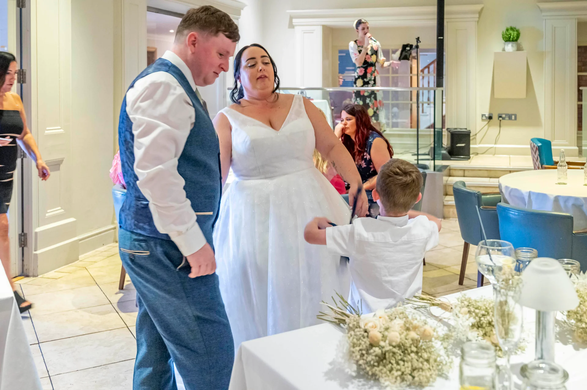 A bride in a white A-line dress and a groom in a blue waistcoat stand together at their wedding reception while a young page boy in a white shirt interacts with them near a decorated table.