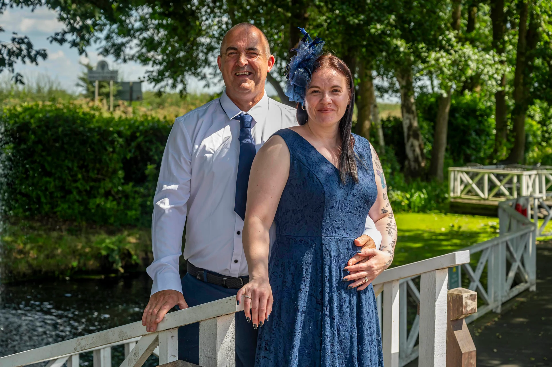 An older couple in formal wedding attire pose together on a white wooden bridge amidst lush greenery and trees on a sunny day.