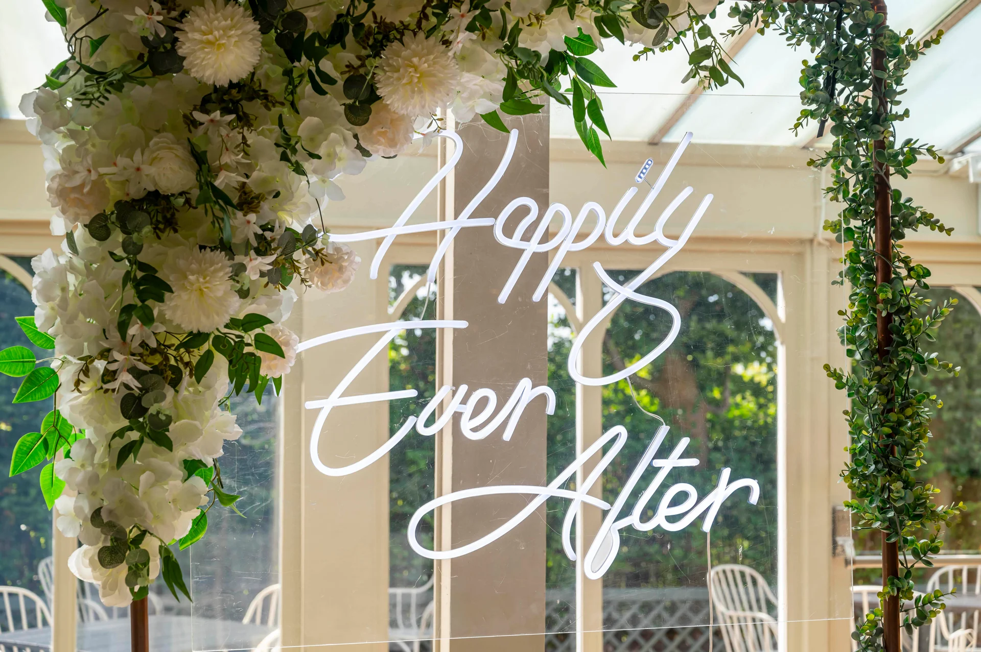 A clear acrylic wedding sign featuring a white neon script that reads Happily Ever After, adorned with lush white floral and eucalyptus arrangements.
