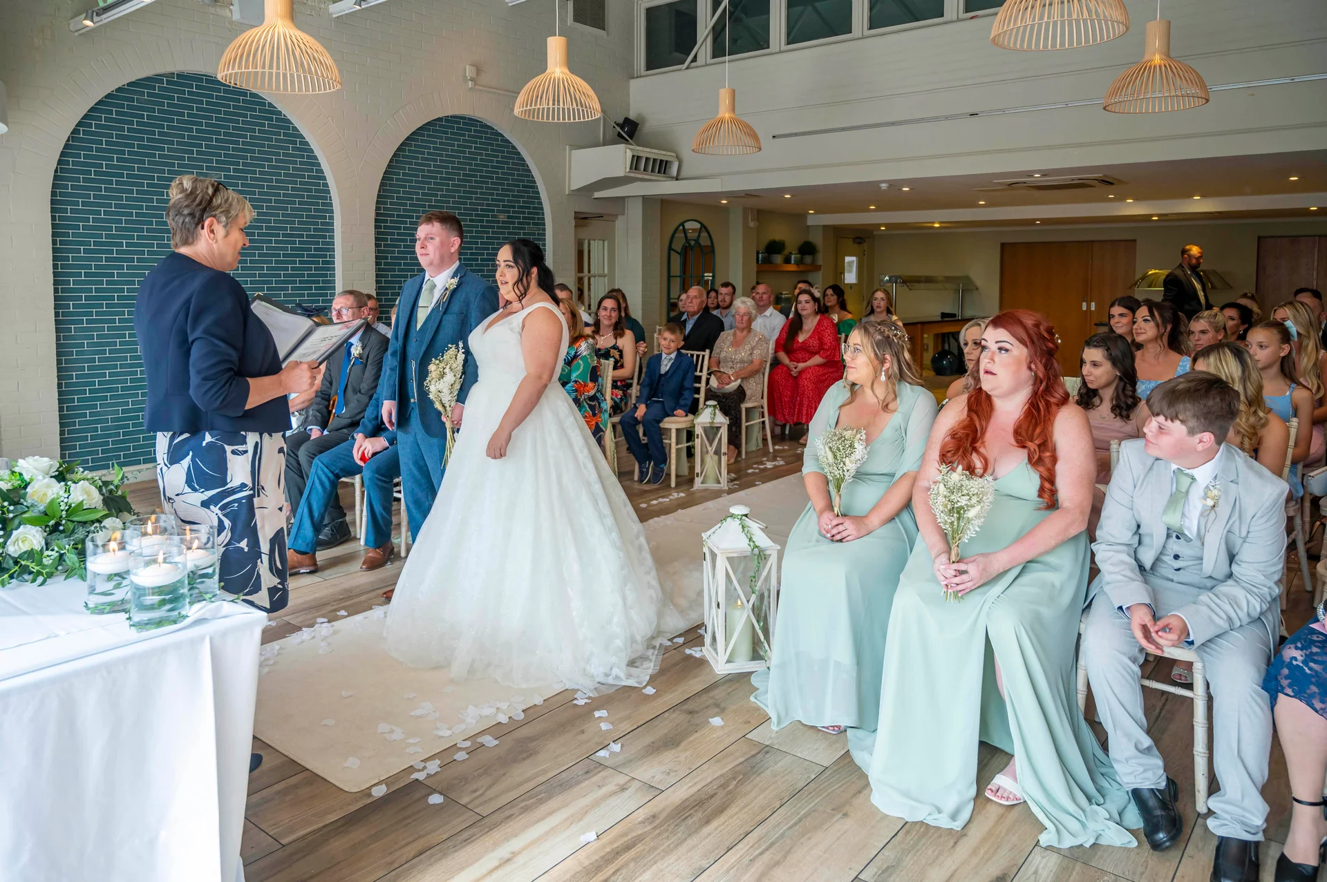 A bride and groom stand at the altar during a civil wedding ceremony surrounded by seated guests, bridesmaids in sage green dresses, and a celebrant.