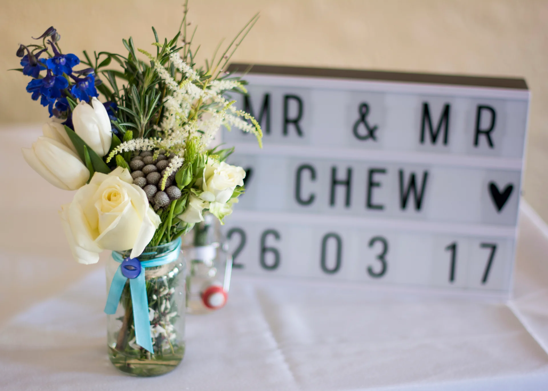 A rustic mason jar filled with white roses, tulips, blue delphiniums and greenery sits beside a lighted sign reading Mr and Mr Chew on a white tablecloth.