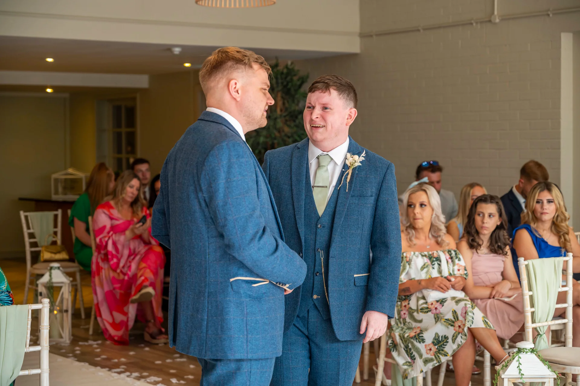 Two grooms in matching blue tweed suits stand together during their wedding ceremony, with one groom looking emotional and smiling as they exchange vows.