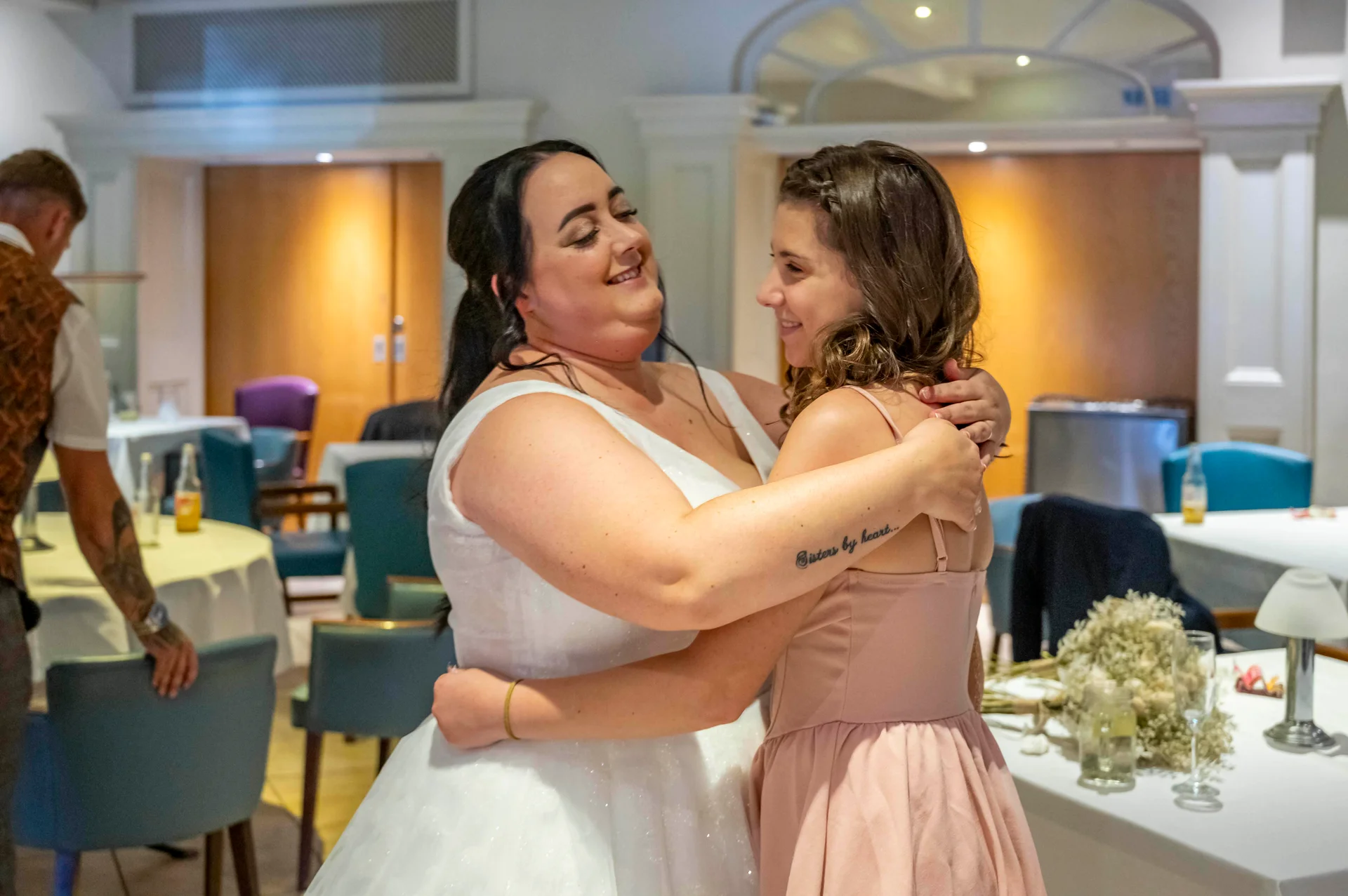 A joyful bride in a white gown shares a warm, laughing embrace with a guest wearing a pink dress during the wedding reception.