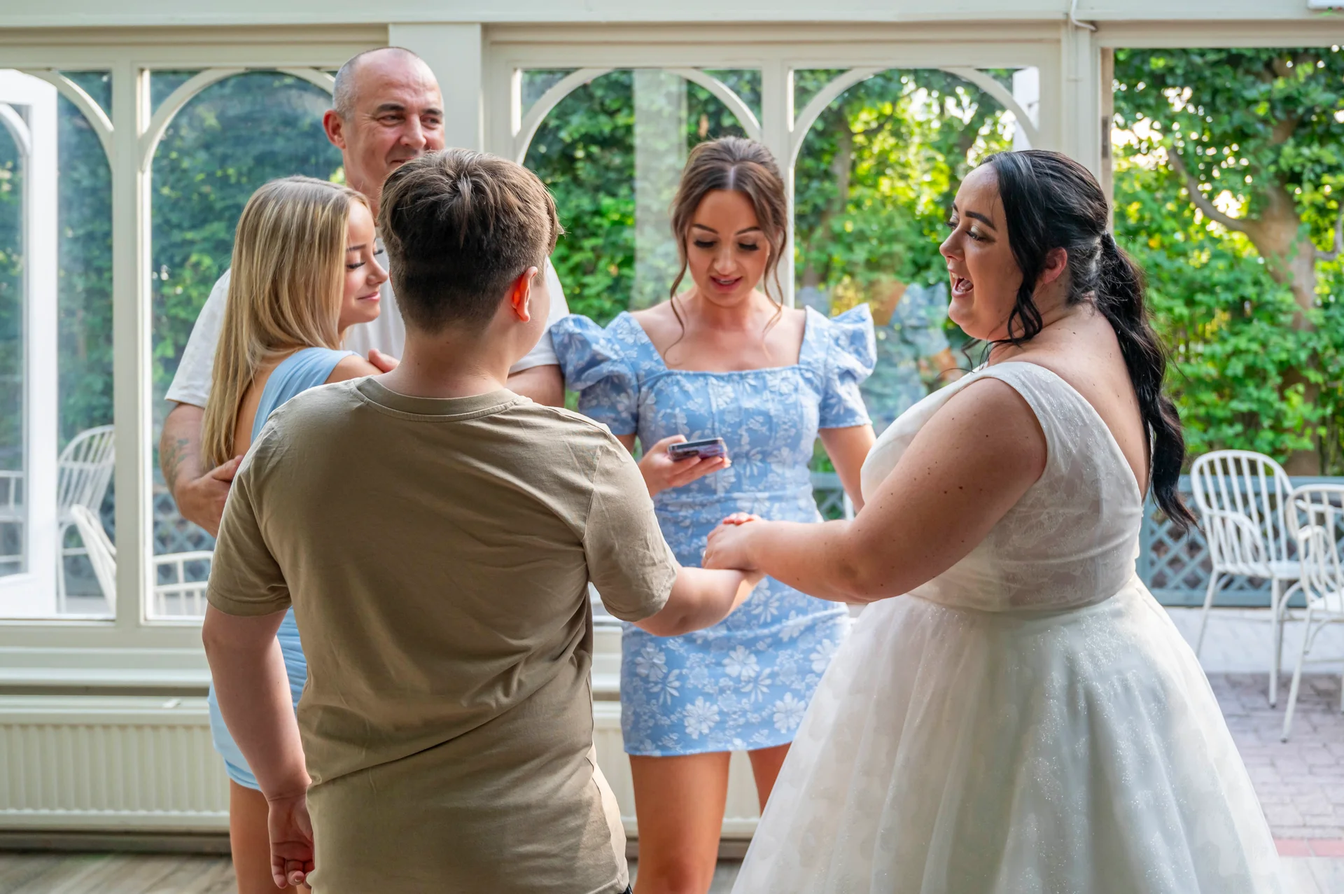 A joyful bride in a white wedding gown holding hands and laughing with family members inside a sunlit conservatory venue.