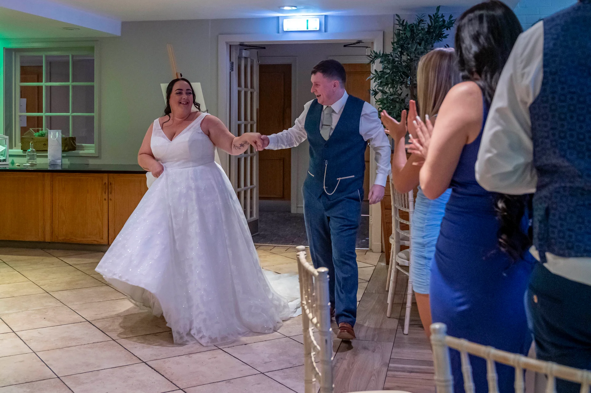A smiling bride in a sparkly a-line gown and a groom in a blue three-piece suit joyfully hold hands as they enter the reception room while guests applaud.