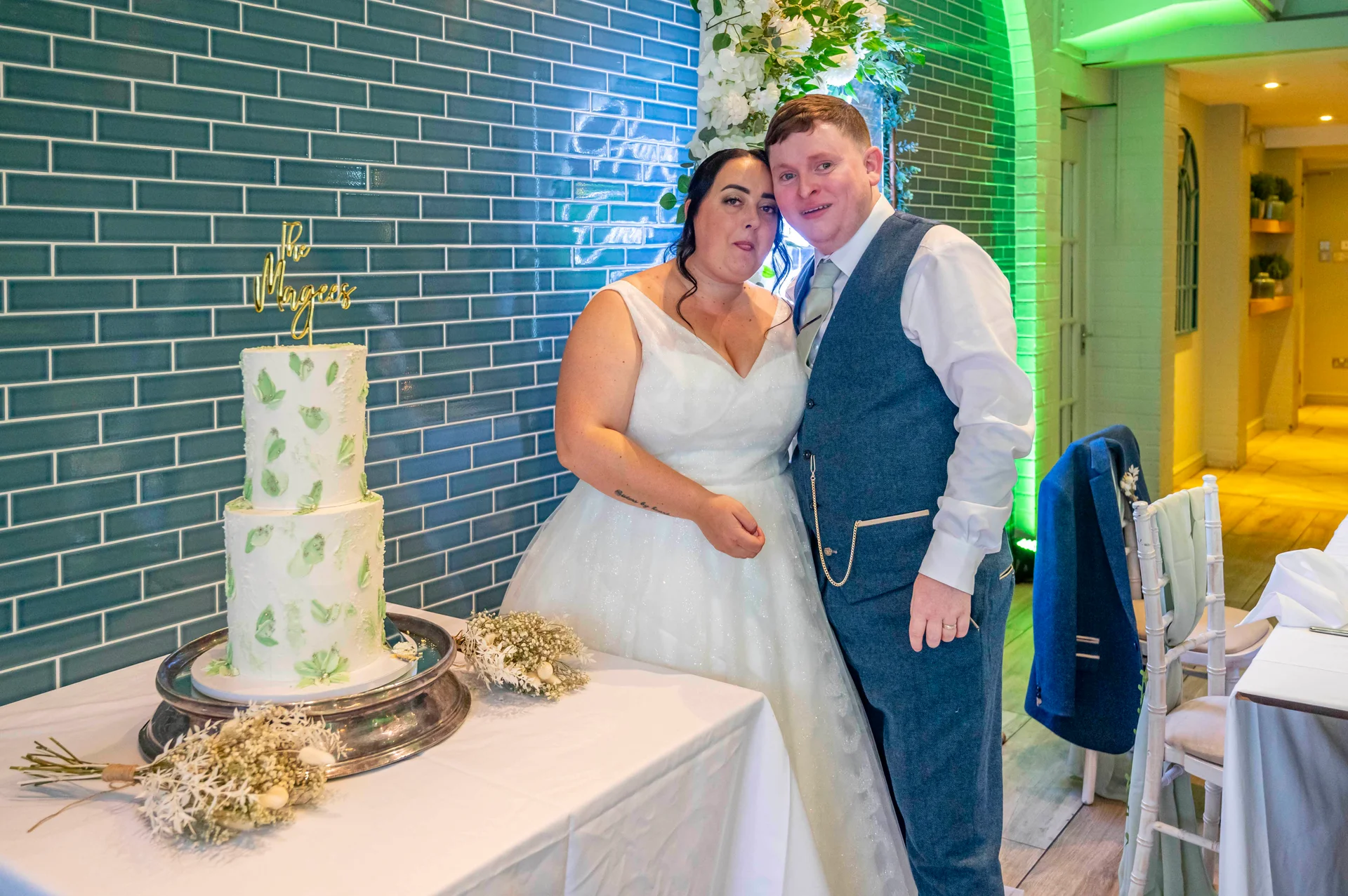 A bride and groom pose smiling next to a two-tier painted wedding cake featuring green leaf motifs against a striking blue tiled wall.