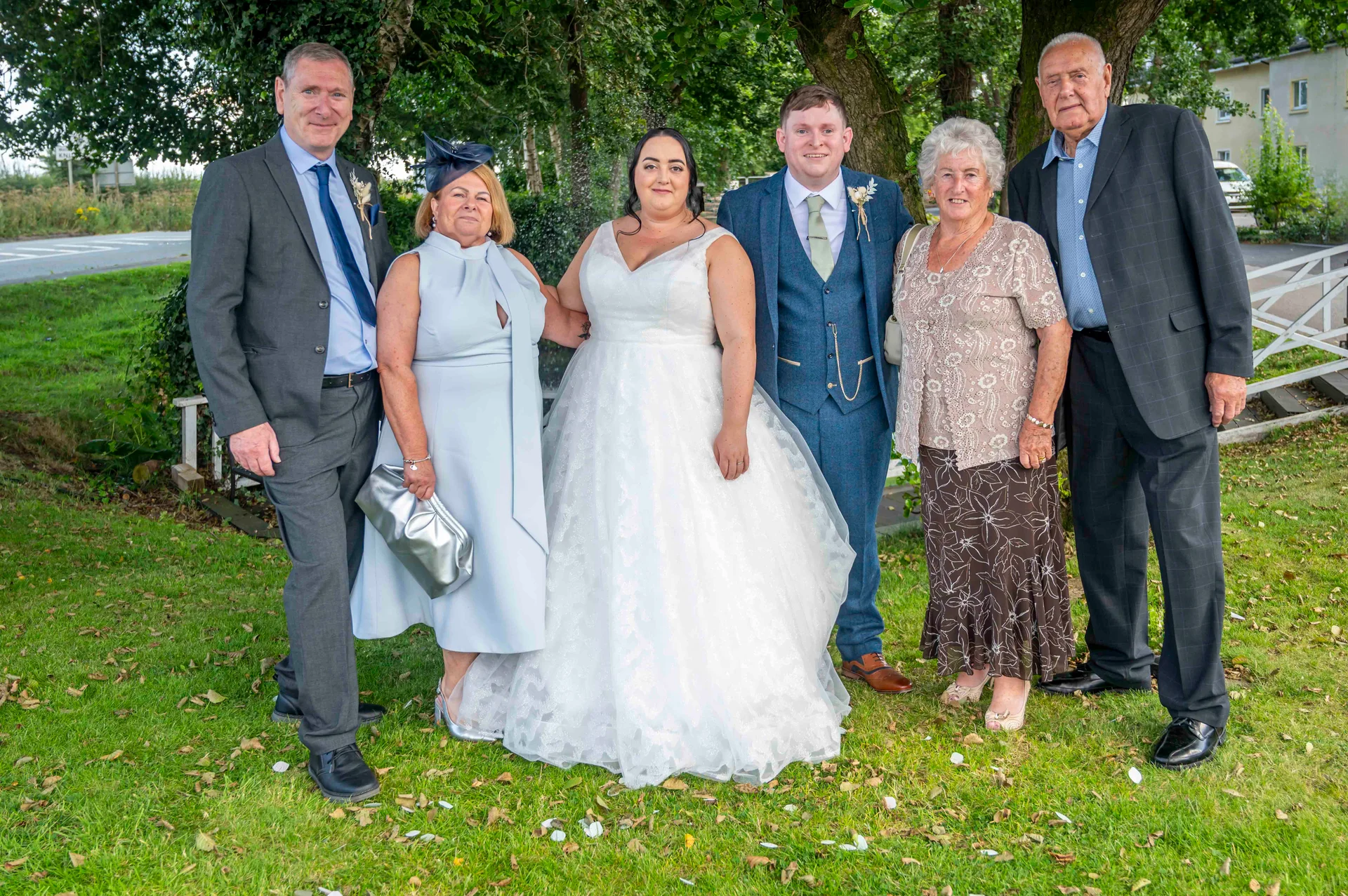A newlywed couple poses outdoors on a grassy lawn with family members, including parents and grandparents, dressed in formal wedding attire.