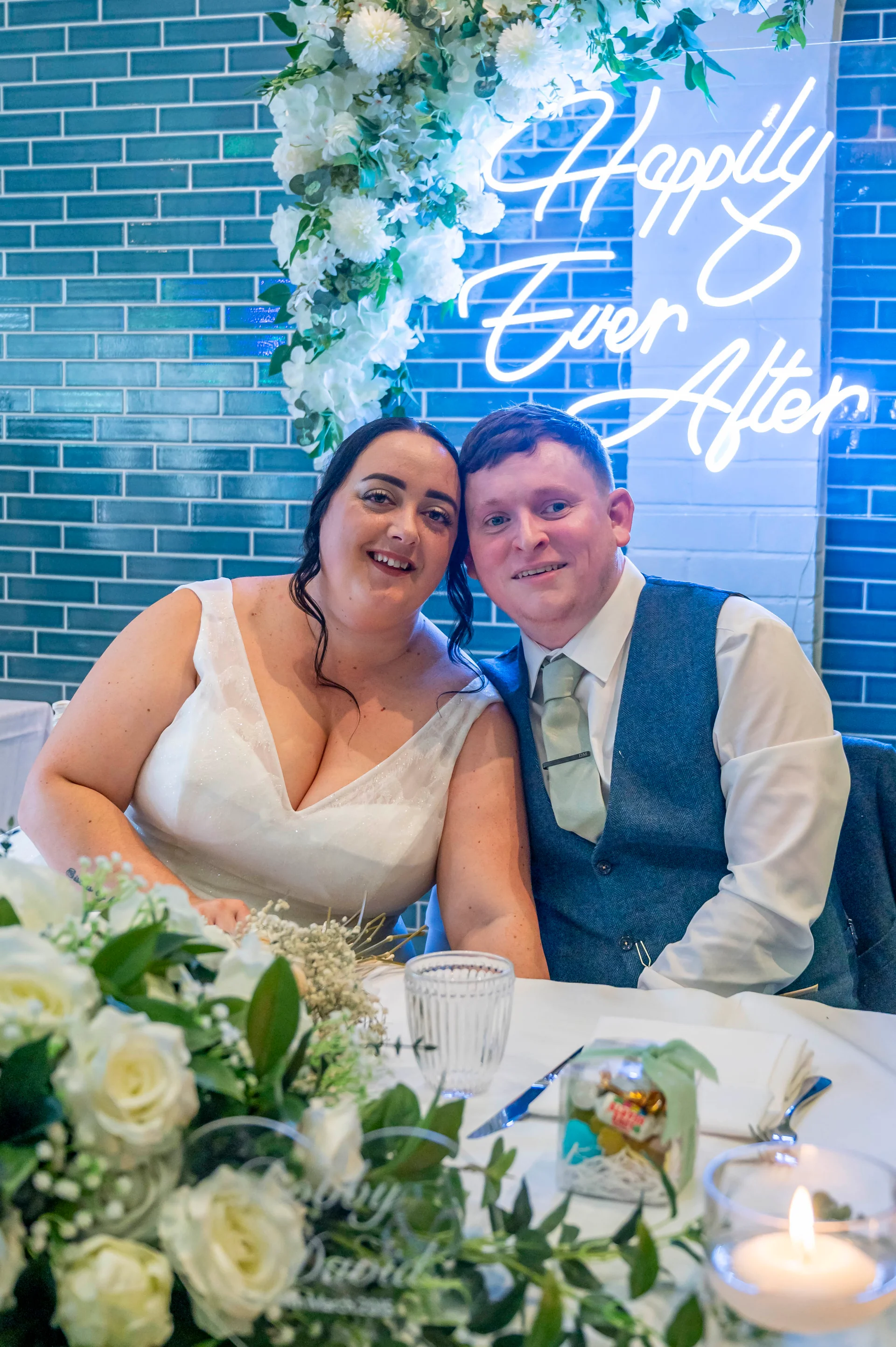 A smiling bride and groom sit together at a top table decorated with white roses and candles, in front of a blue tiled wall and a bright neon sign.