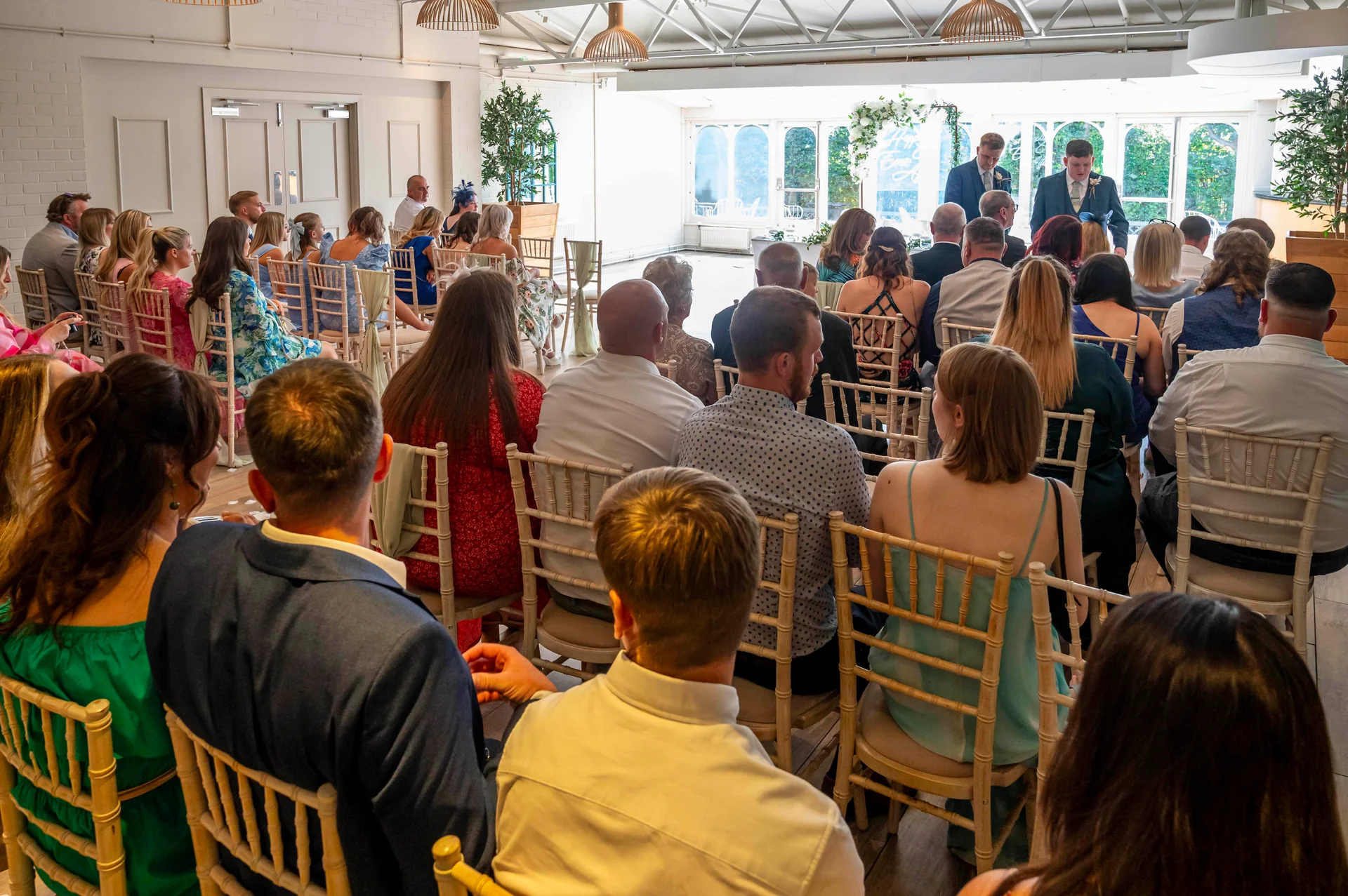 Guests are seated inside a bright orangery facing two grooms standing at the altar during a wedding ceremony.