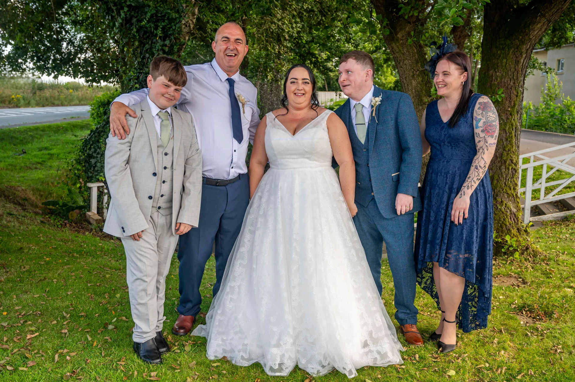 A happy wedding group stands outdoors on a grassy lawn next to a mature tree, featuring a smiling bride in an A-line gown, a groom in a blue tweed suit, and family members including a page boy and a guest in a navy dress.