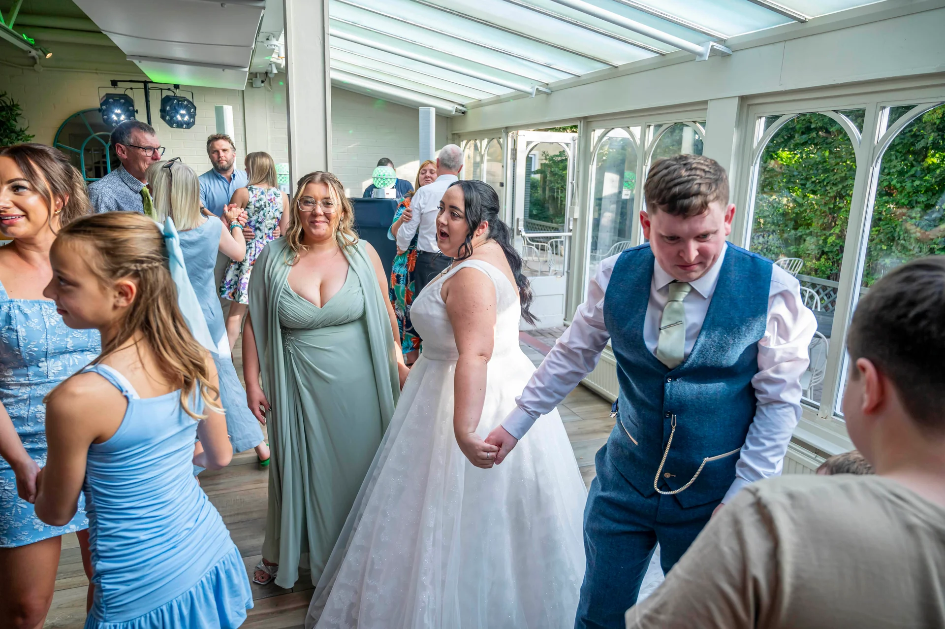 A newlywed couple holds hands and leads guests in dancing on the wooden floor of a light-filled conservatory wedding venue.
