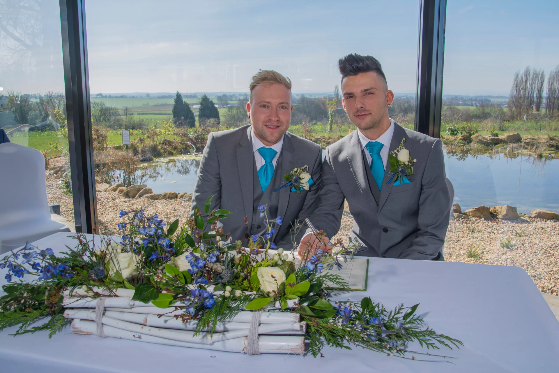 Two grooms in matching grey suits and bright blue ties smile as they sign the wedding register at a table adorned with a blue and white floral arrangement against a scenic window backdrop.