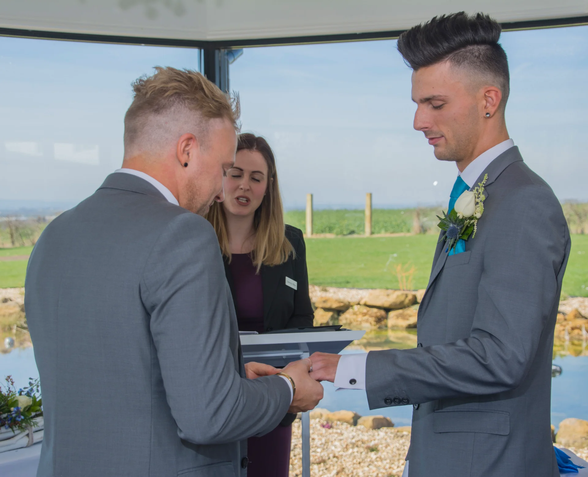 Two grooms in matching grey suits exchange wedding rings during an outdoor civil ceremony officiated by a celebrant.