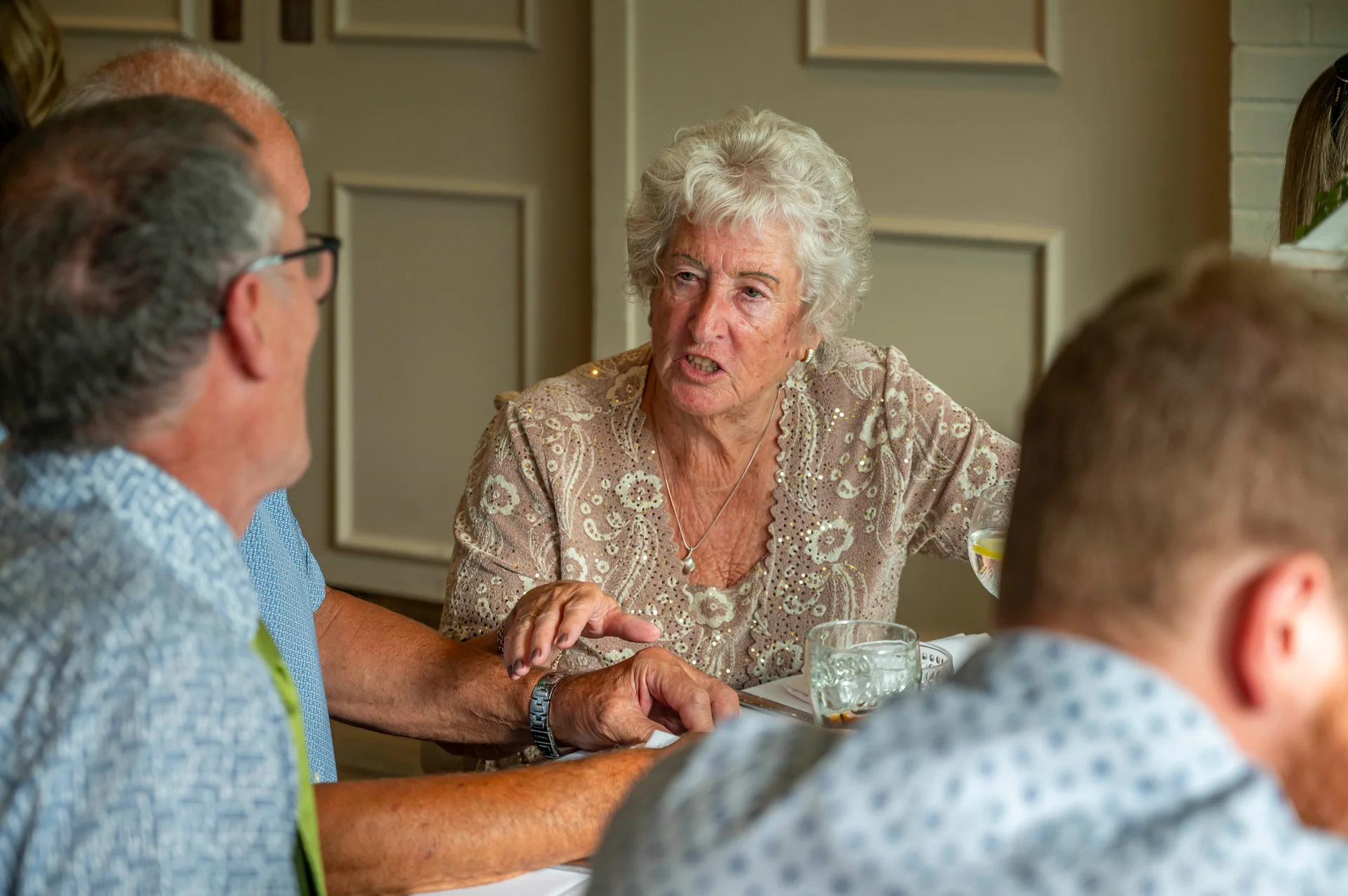 An older woman with short curly grey hair speaks animatedly across a wedding reception table while guests chat around her.