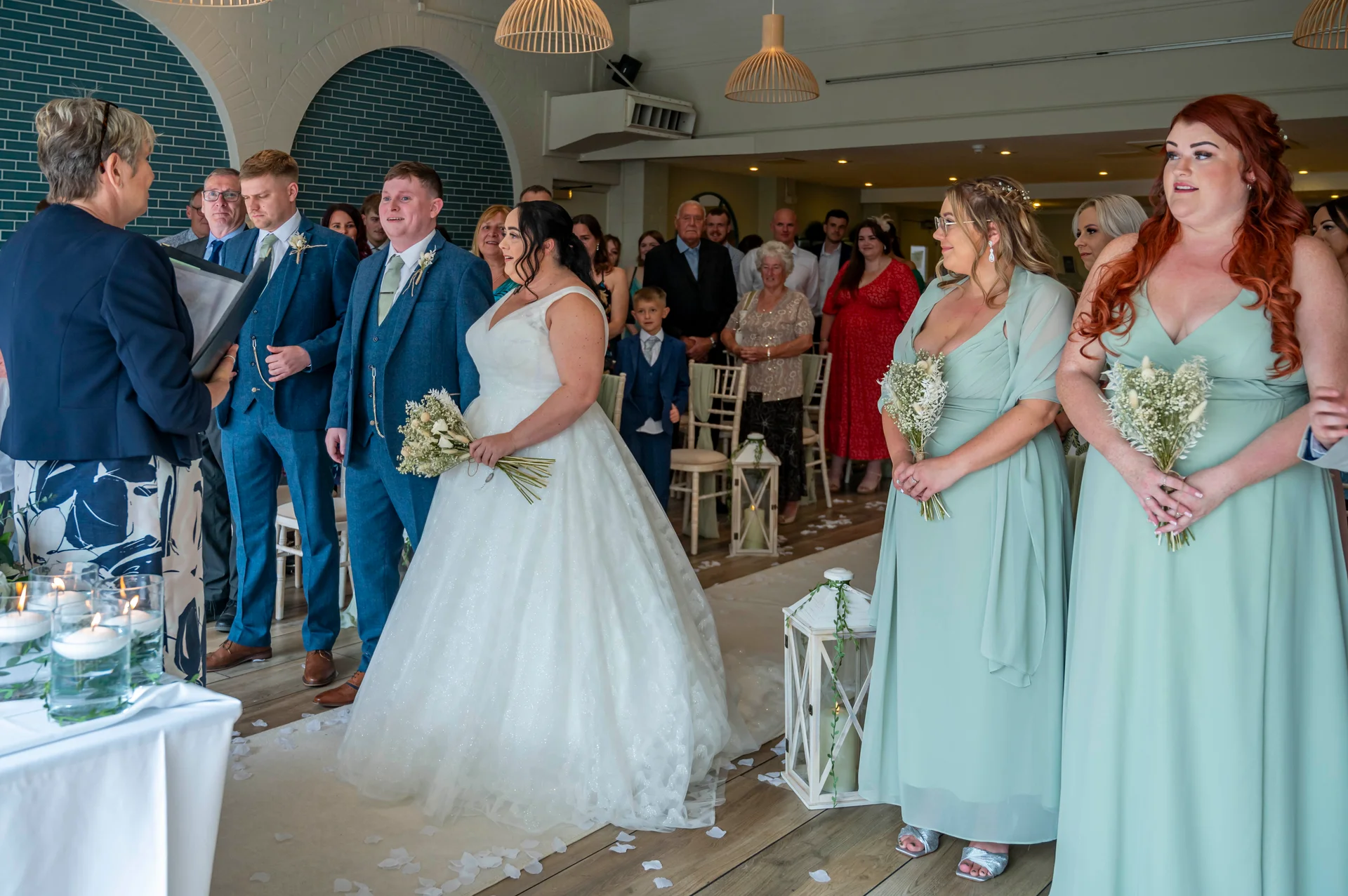 A bride in a white gown and groom in a blue suit stand during a civil ceremony with bridesmaids in sage green dresses holding bouquets.