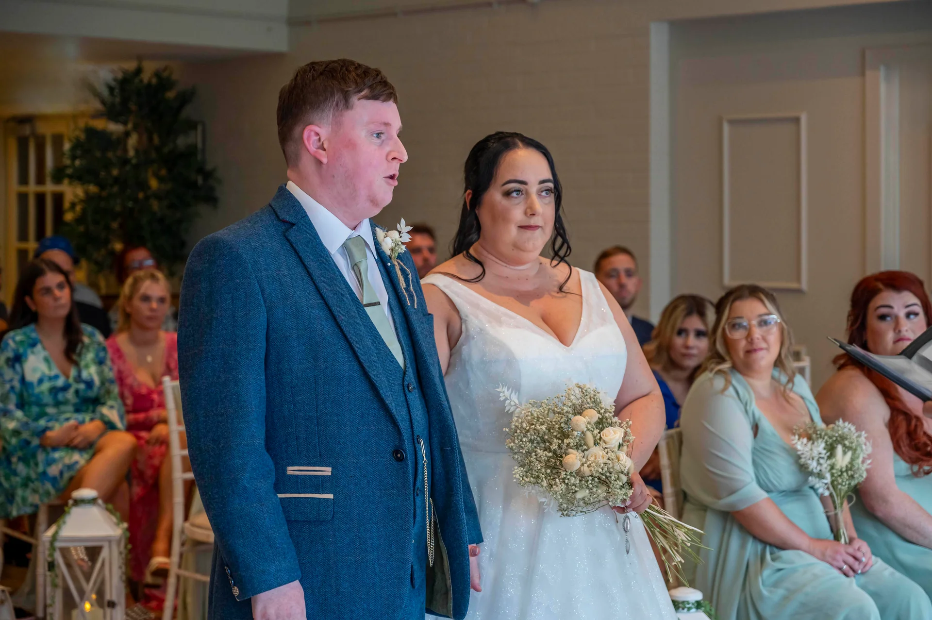A groom in a blue suit and a bride in a white gown stand side by side during their indoor wedding ceremony, surrounded by seated guests and bridesmaids holding small bouquets.