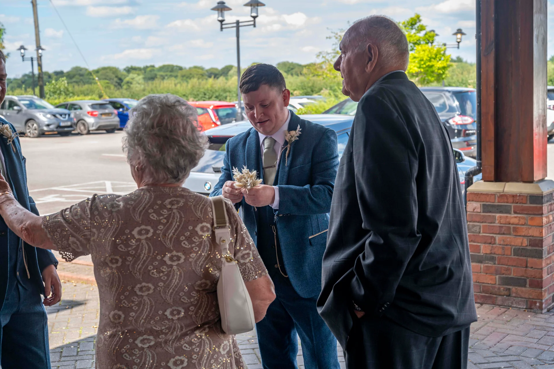 A groom in a blue tweed suit stands outdoors in a car park chatting and looking at small floral buttonholes with older guests during the wedding day.