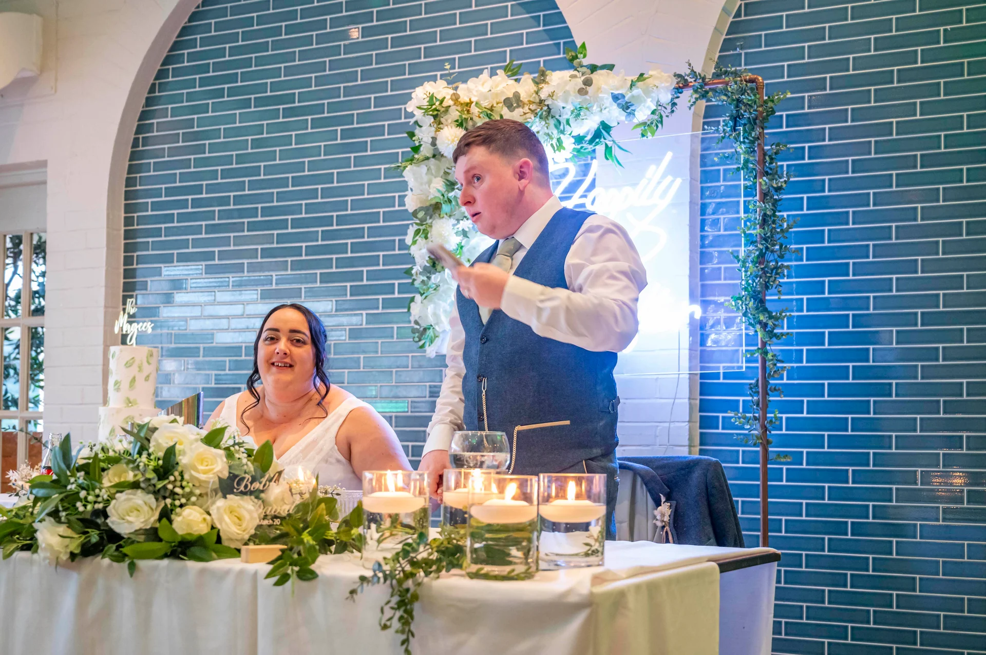 A groom delivers his speech at the top table while his partner looks on smiling, in front of a blue tiled feature wall and illuminated neon backdrop.