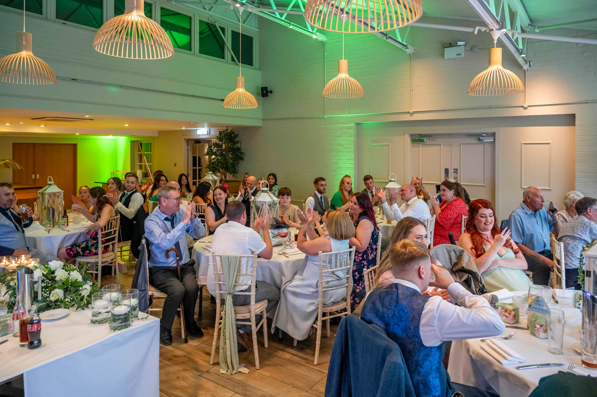 Wedding guests seated at round tables cheer and clap during the wedding breakfast in a spacious warehouse venue with vaulted ceilings and hanging pendant lights.