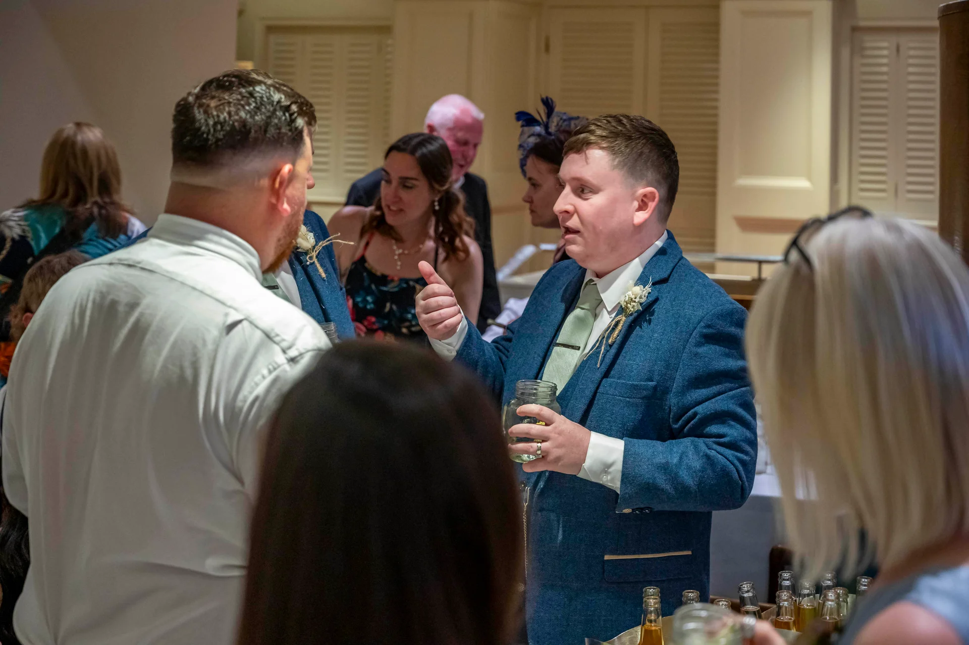 Wedding guests mingling and chatting during the drinks reception, with a groom in a blue tweed suit holding a drink.