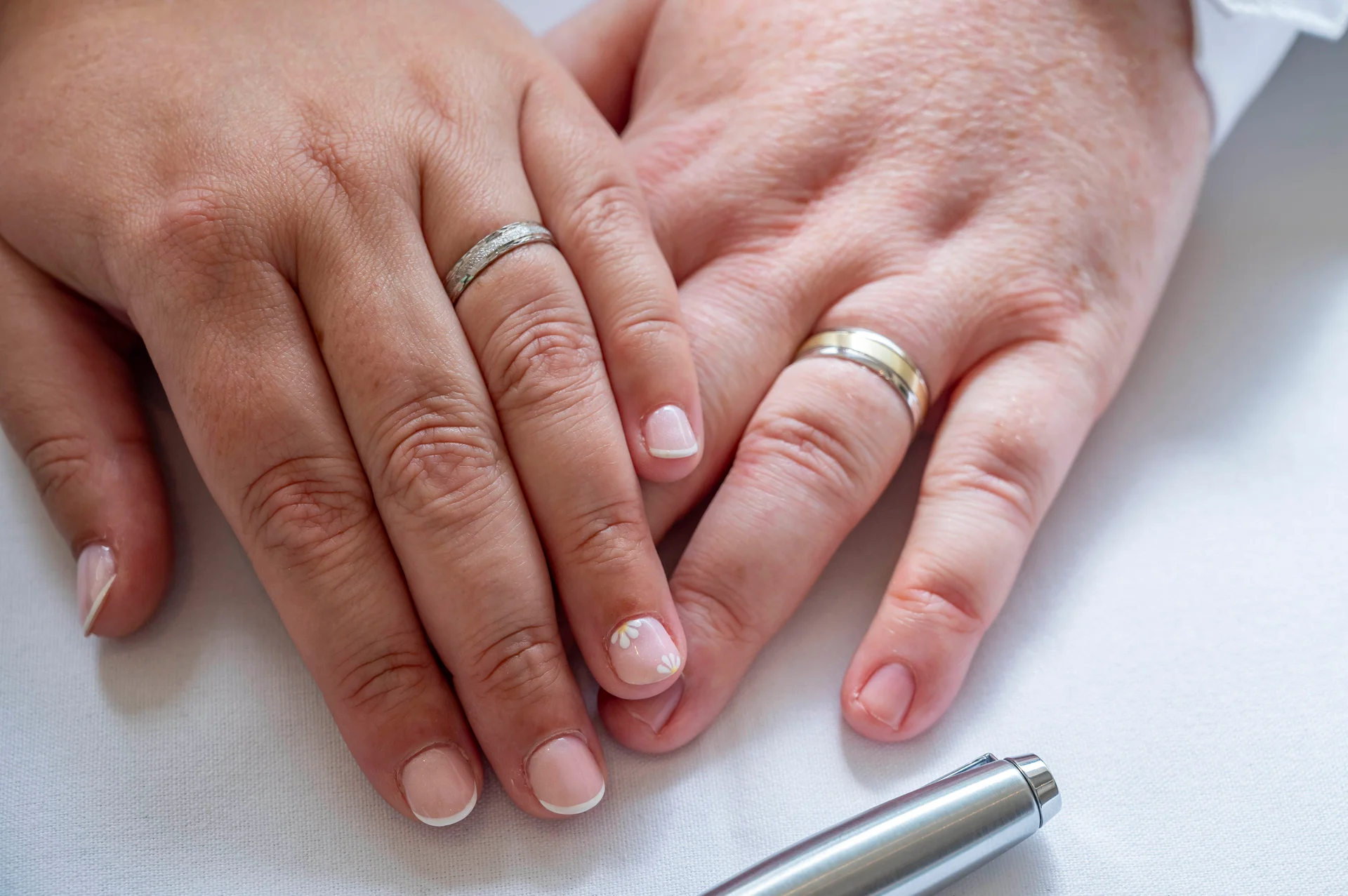 Close-up shot of an older couple's hands resting together on a white surface, showcasing their newly exchanged wedding bands and manicured nails.