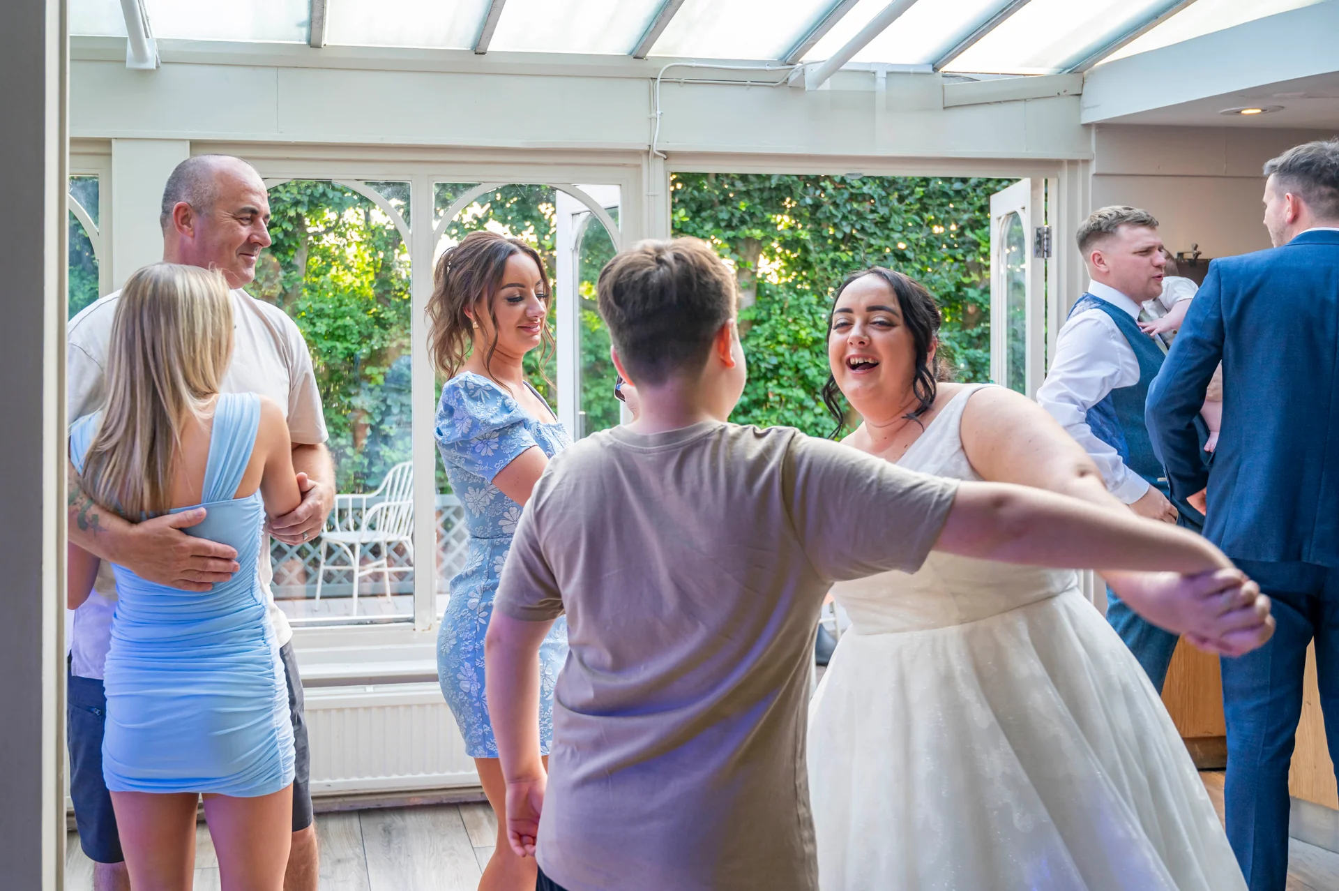 A joyful wedding guest laughs while dancing with the bride inside a bright conservatory filled with natural light.