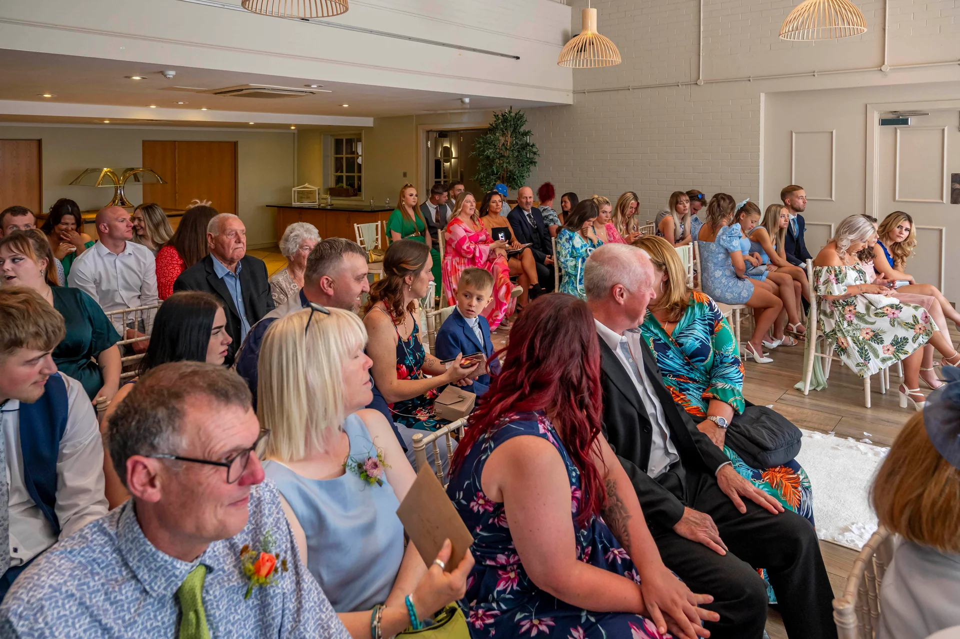 A candid wide shot captures a packed room of wedding guests smiling and chatting during a ceremony in a bright, modern venue.