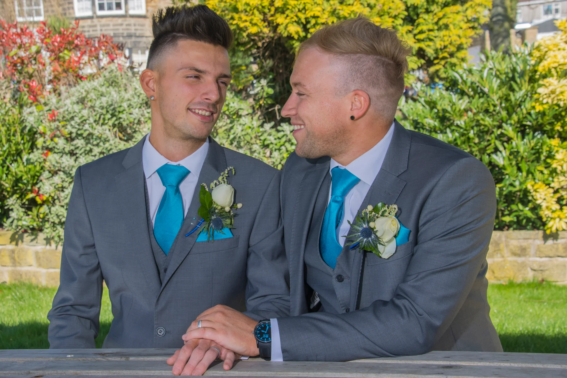 Two grooms in matching grey suits and vibrant turquoise ties exchange a loving glance outdoors with white floral buttonholes.