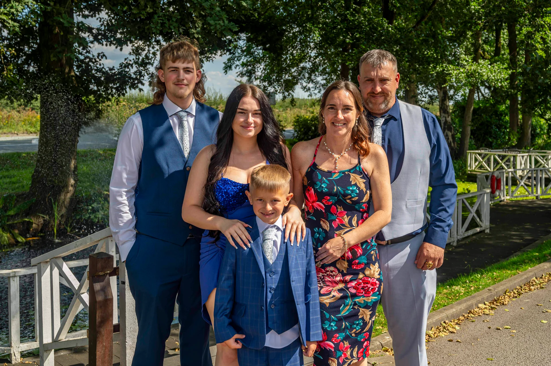 A formal family group poses outdoors beside a white wooden bridge on a sunny wedding day, wearing coordinating blue and grey suits alongside floral guest attire.