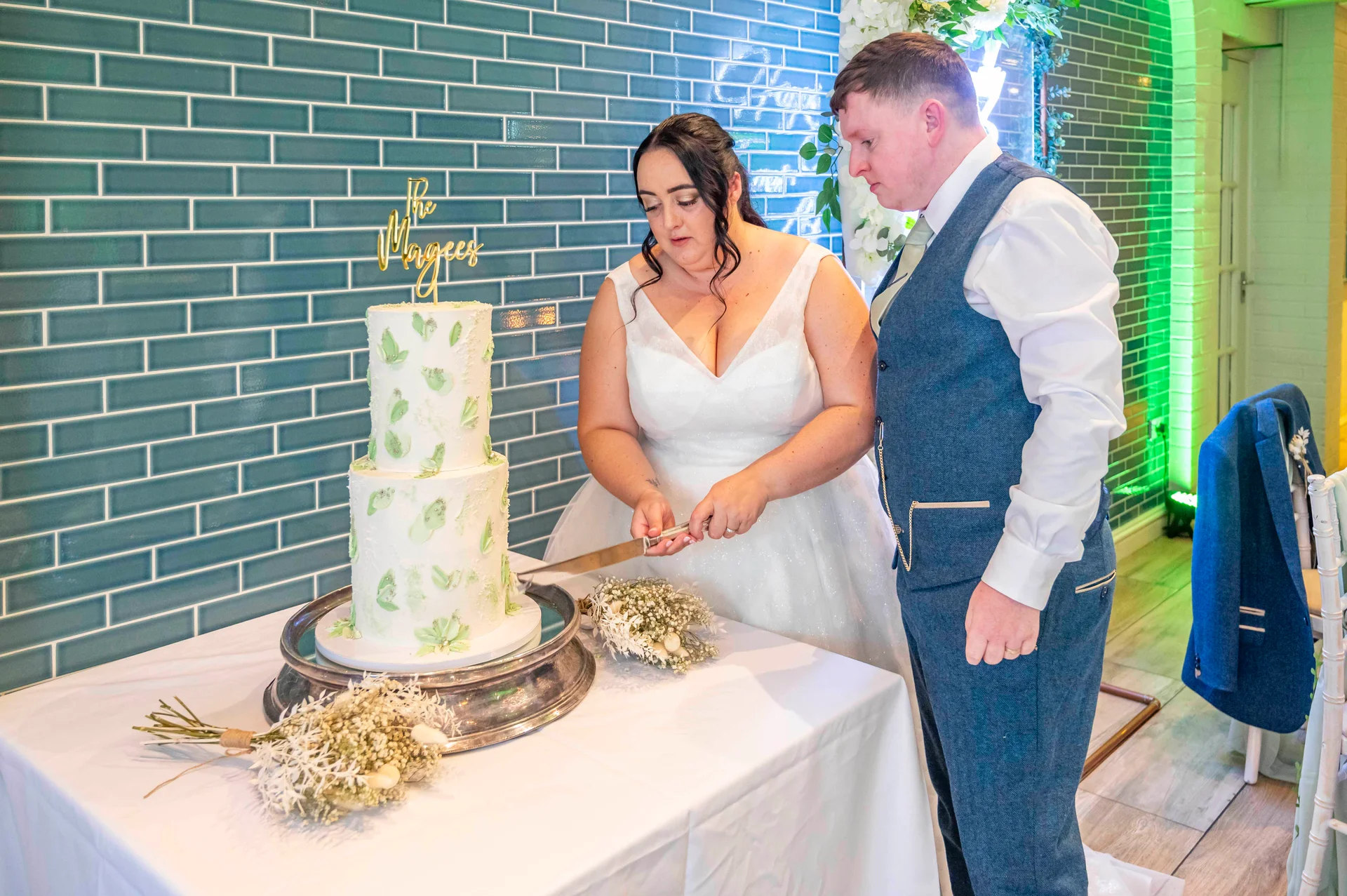 A newlywed bride and groom cut a two-tiered white cake with green leaf details against a blue tiled wall.