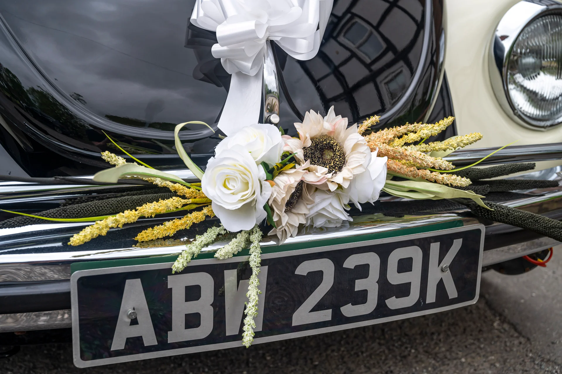 Close-up of a classic vintage Volkswagen Beetle wedding car decorated with a white ribbon bow and an arrangement of white roses and yellow florals across the chrome bumper.