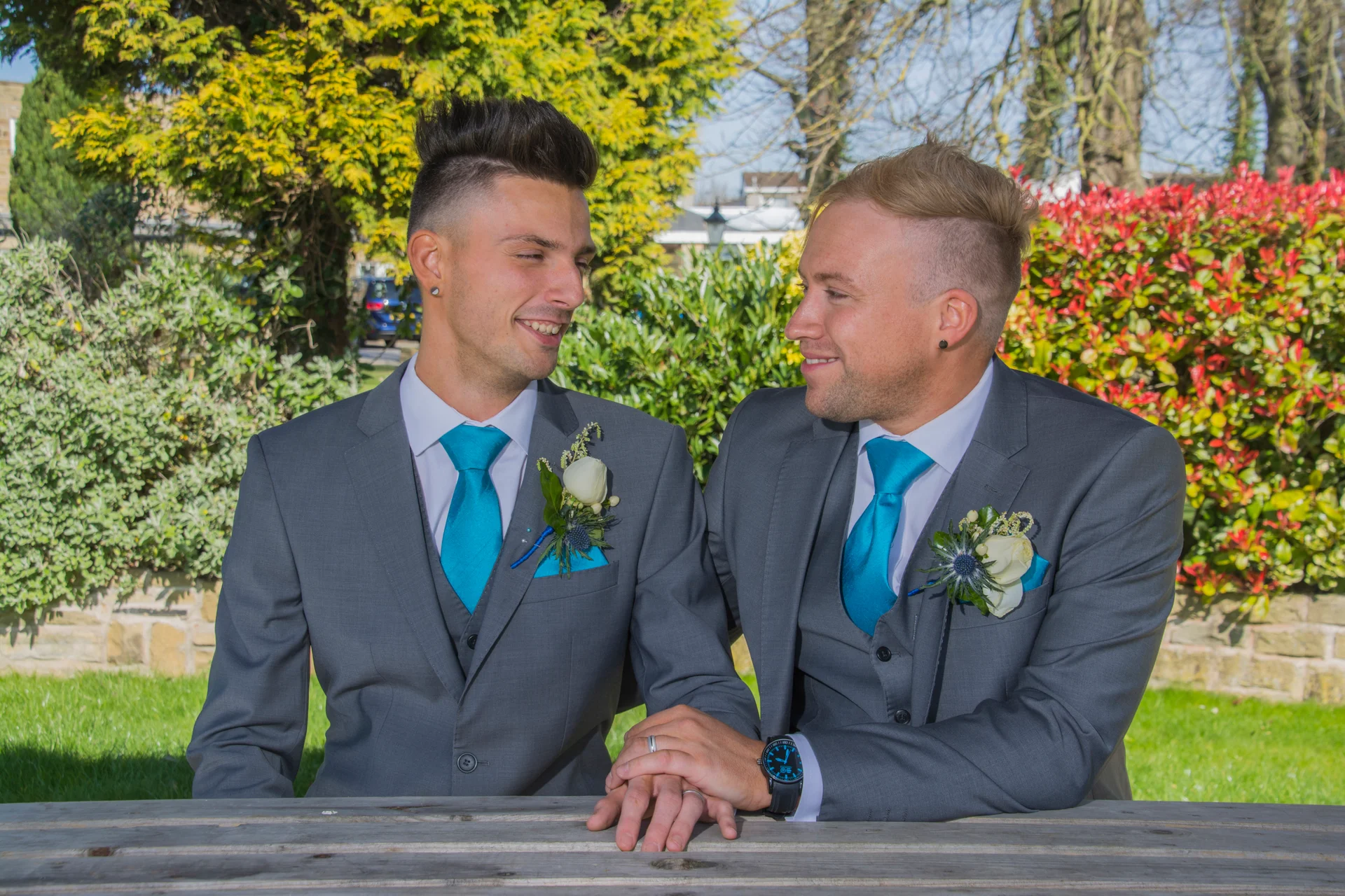 Two grooms in matching grey three-piece suits with bright blue ties and white rose buttonholes share a loving glance while holding hands outdoors in a sunny garden.