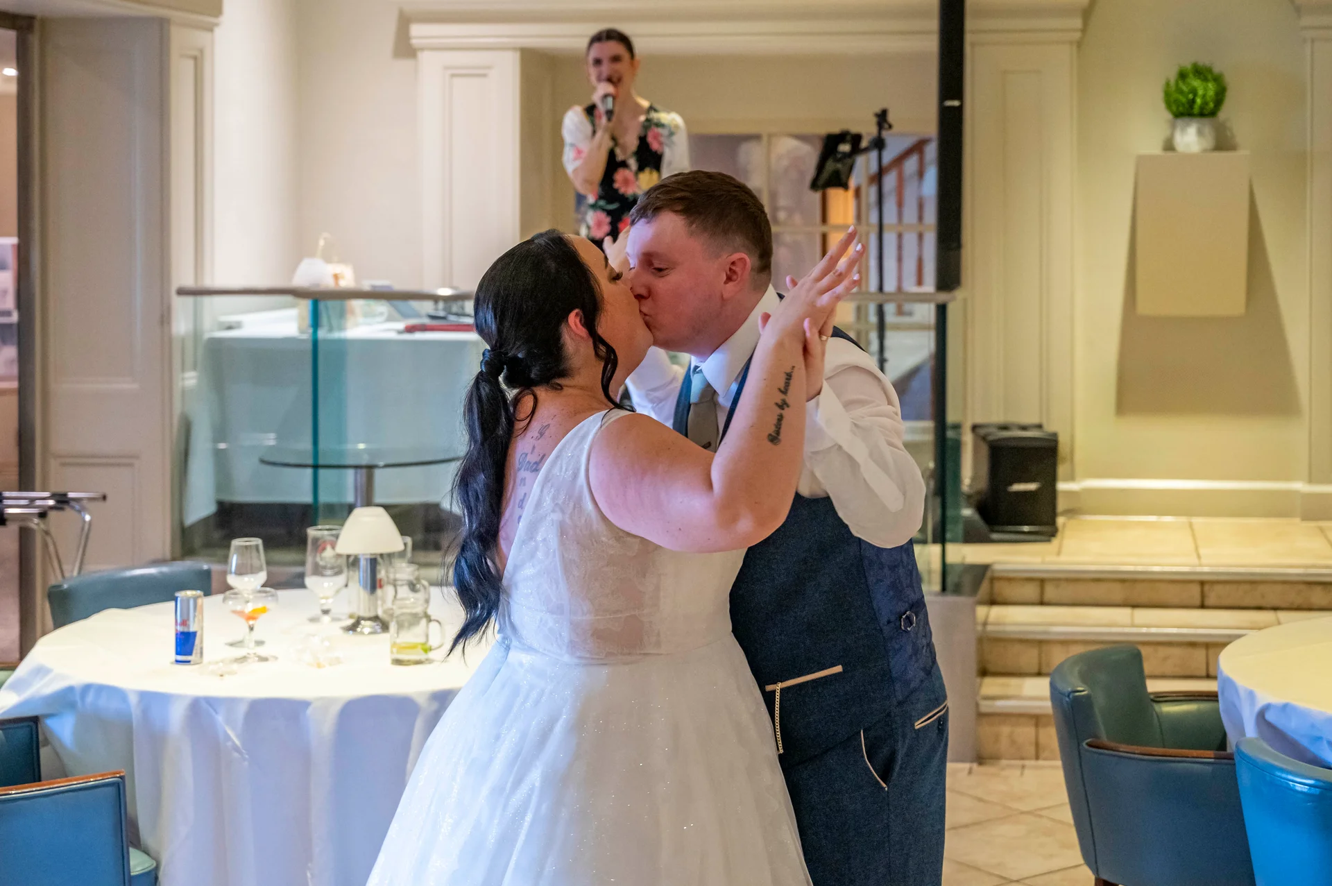 A newlywed couple share a tender kiss during their first dance indoors, with a singer performing in the background near a set of dining tables.