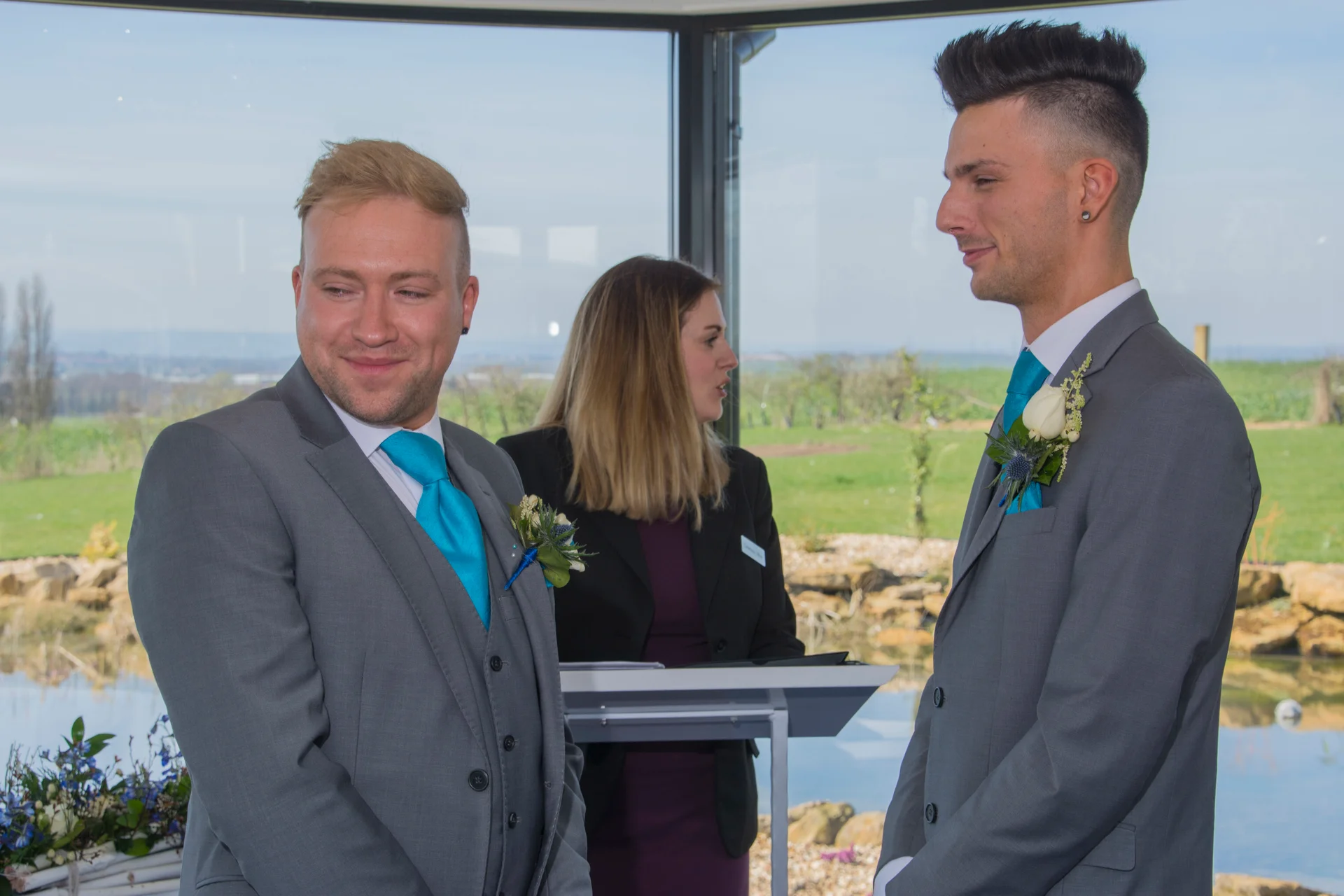 Two grooms in matching grey suits and bright blue ties share a joyful moment during their wedding ceremony.