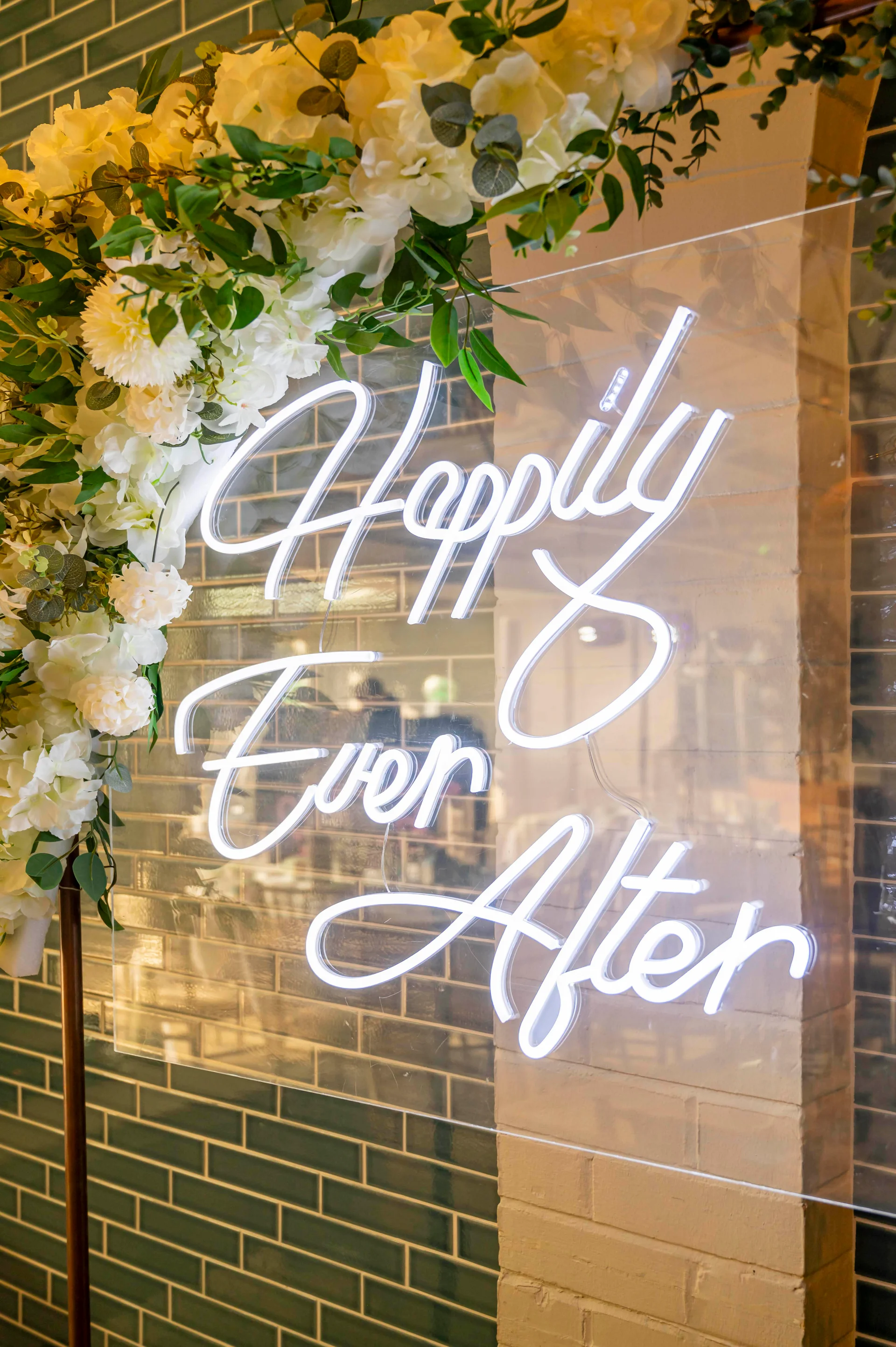 A glowing white neon sign that reads happily ever after, mounted on clear acrylic and decorated with a lush arrangement of white flowers and greenery against an exposed brick wall.