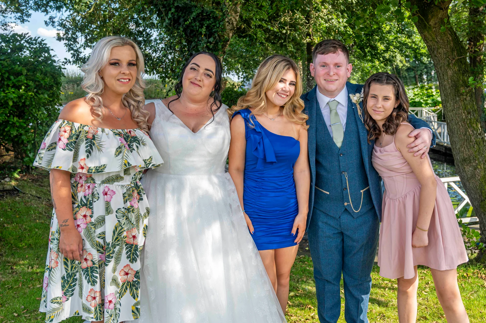 A smiling wedding party poses outdoors in a sunlit garden, featuring the bride in a white gown and the groom in a blue tweed suit alongside guests in floral and colourful dresses.