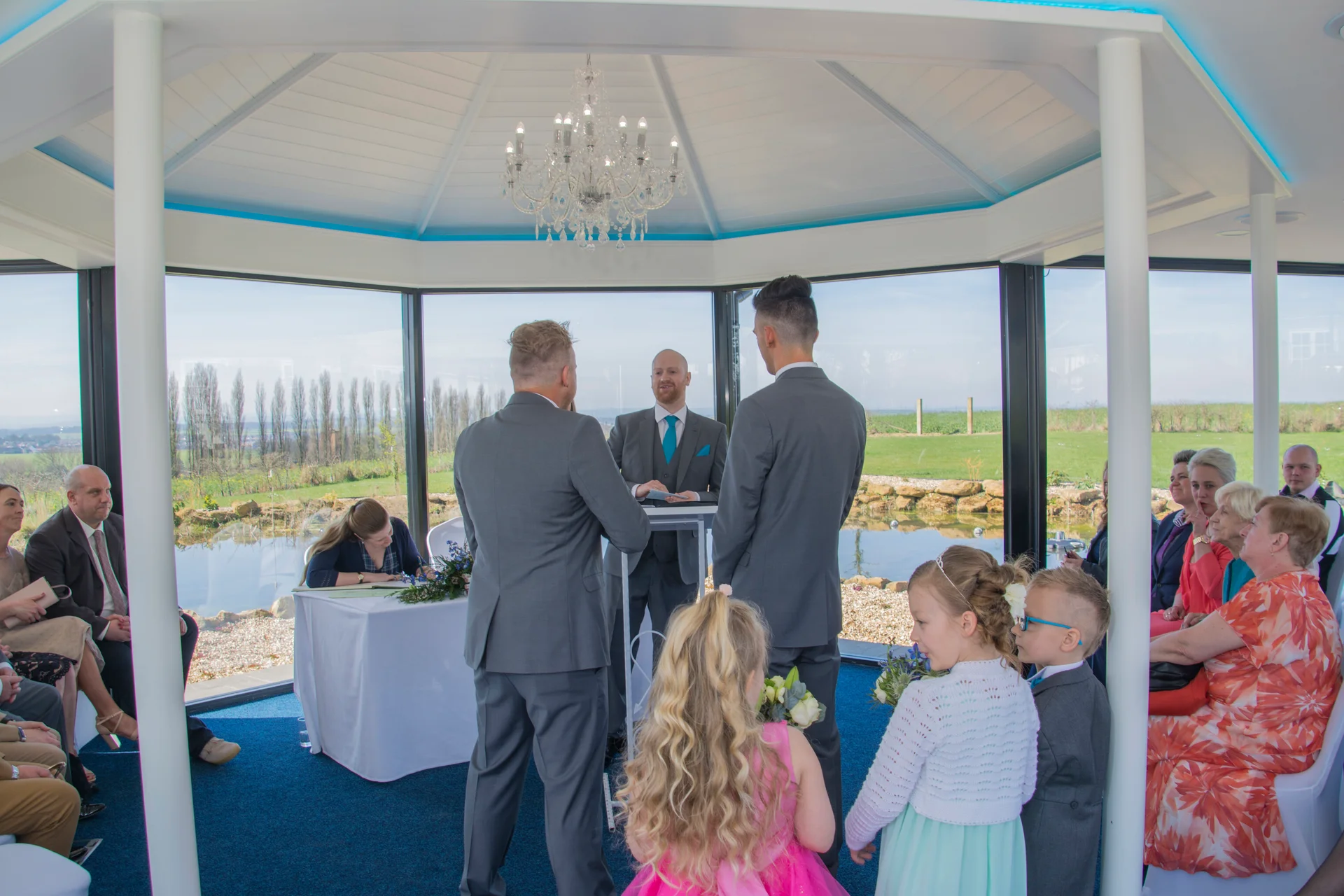 A same-sex couple exchanges vows during a light-filled indoor ceremony surrounded by panoramic windows, family members, and a wedding party.