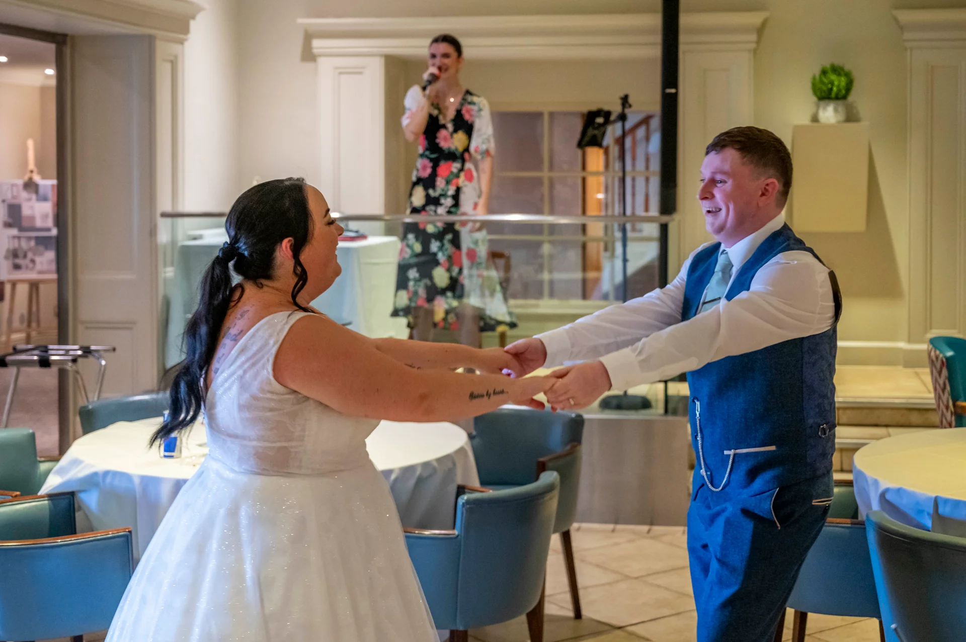 A newlywed couple laugh and hold hands while sharing their first dance in a hotel function room as a singer performs in the background.