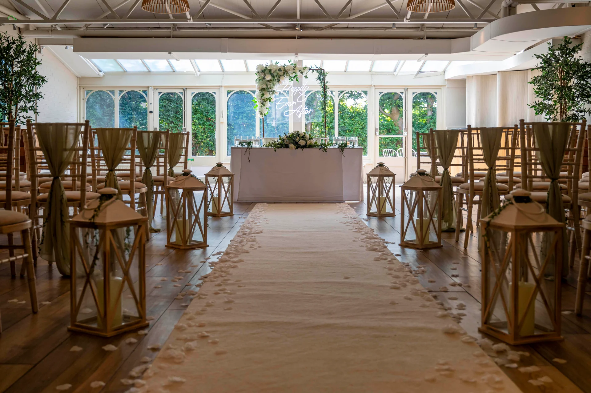 An elegantly styled indoor wedding ceremony aisle is flanked by wooden lanterns and chairs adorned with fabric sashes, leading towards a flower-decorated altar table.