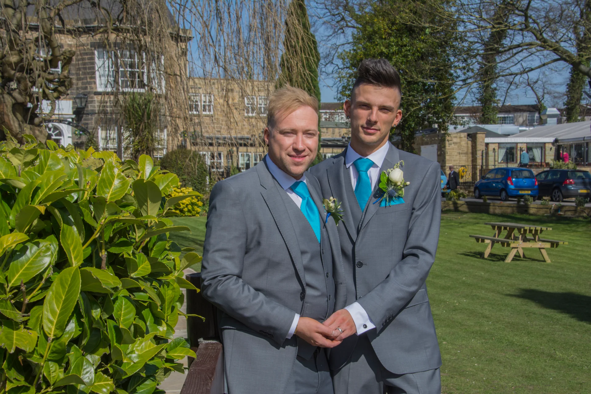 Two grooms stand hand in hand outdoors in matching grey three-piece suits with bright blue ties and boutonnieres in front of a manor house.