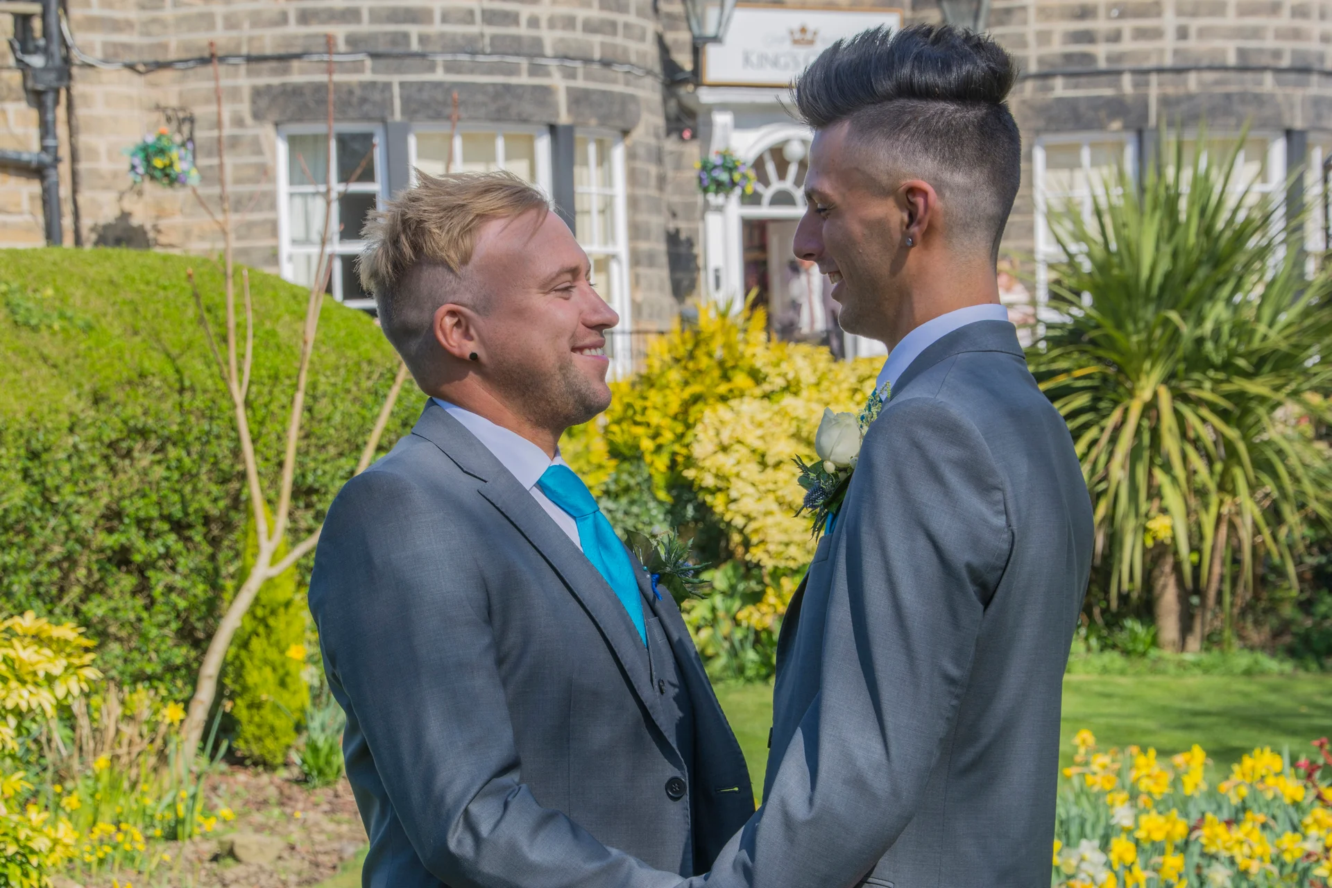 Two grooms in matching grey suits and bright blue ties lovingly look at each other in a sunny hotel garden during their wedding day.