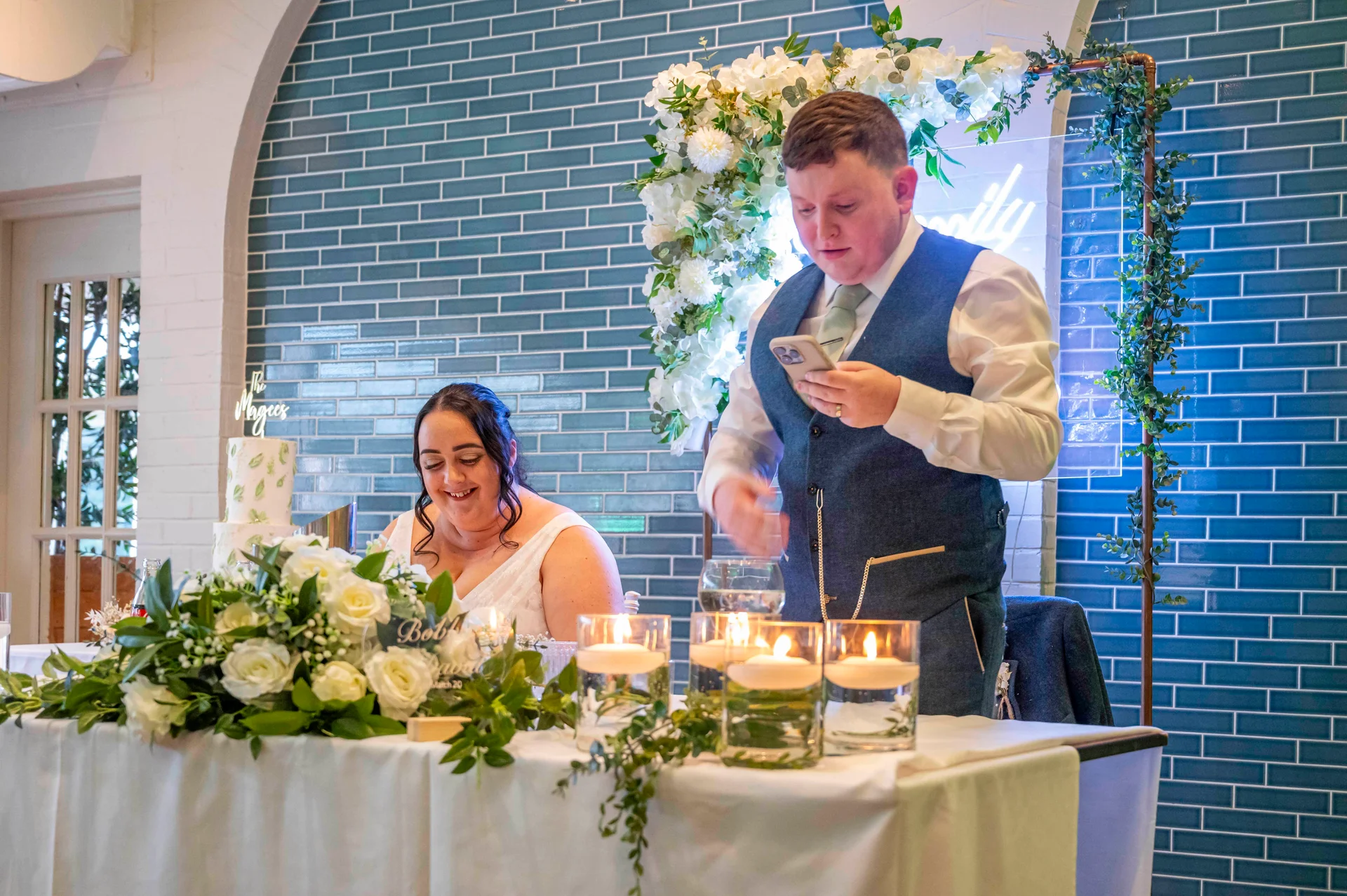 A groom delivers a speech while holding his phone at the top table, seated next to the smiling bride beside a glowing floral backdrop.