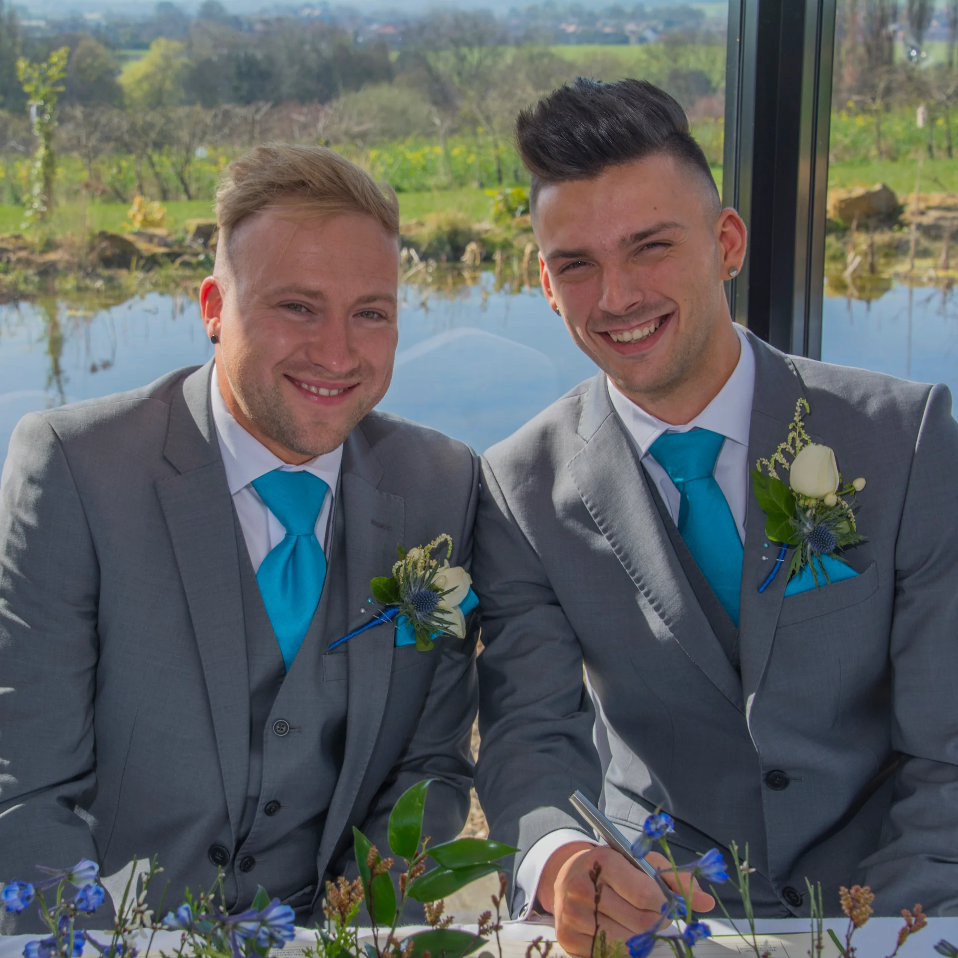 Two smiling grooms in matching grey three-piece suits and bright blue ties sign the register together indoors by a large window overlooking a scenic lake.