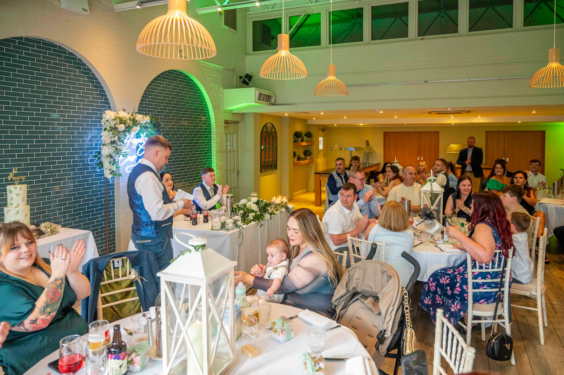 A groom delivers a speech during a wedding reception in a modern restaurant venue with teal tiled walls and pendant lighting, as guests applaud and laugh.