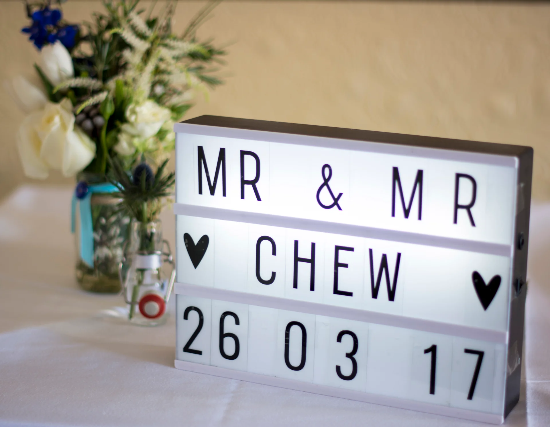 An illuminated lightbox on a table reads MR AND MR CHEW 26 03 17, accompanied by fresh floral arrangements in glass jars.