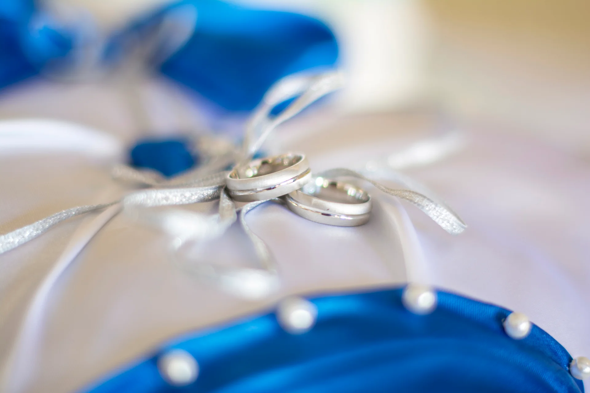 A close-up macro shot displays two silver wedding bands resting delicately on a white and blue decorative ring pillow.