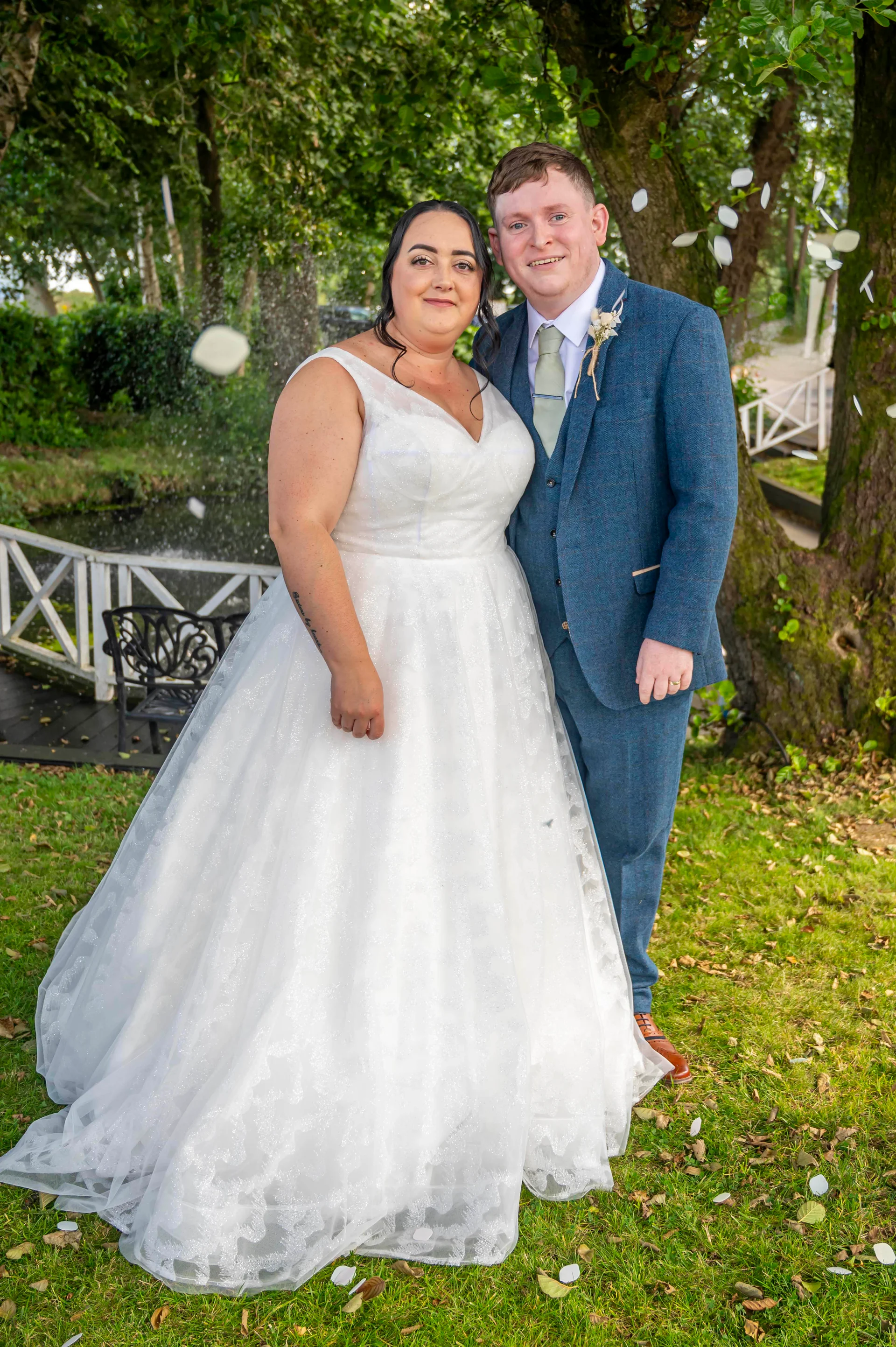 A newlywed couple poses together in a lush garden setting with confetti falling around them, the bride wearing a white a-line wedding dress and the groom in a blue tweed suit.