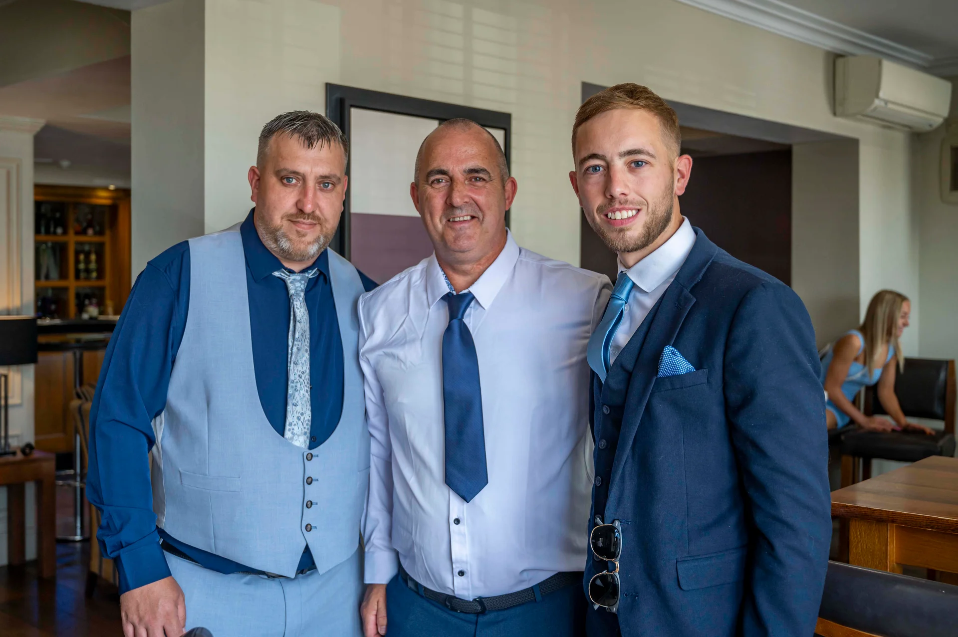 Three smiling groomsmen pose together in matching suits inside a hotel reception venue, with one wearing a blue suit and others in waistcoats.